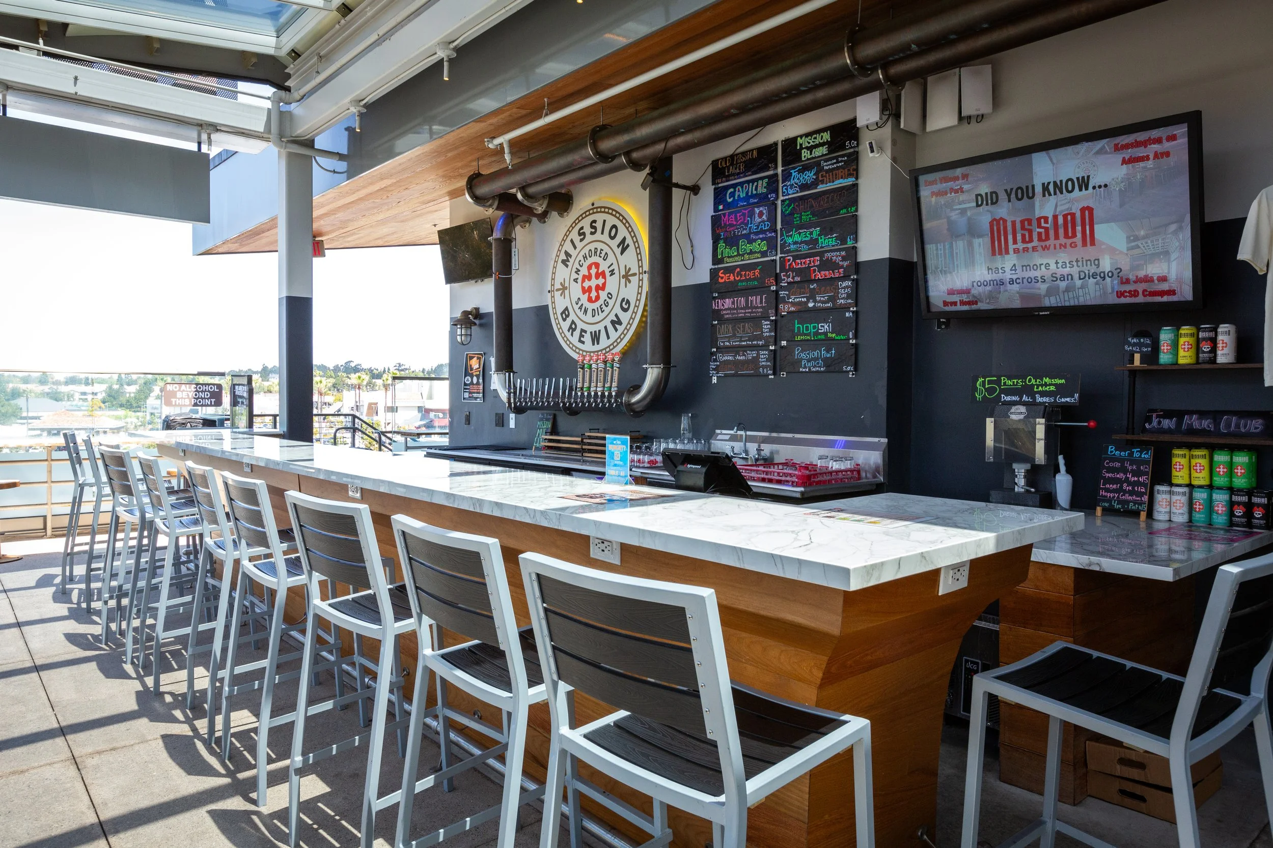 Rooftop bar with a white marble countertop, white chairs, a Mission Brewing sign, a chalkboard menu showing various craft beers, and a large digital screen displaying advertising content.