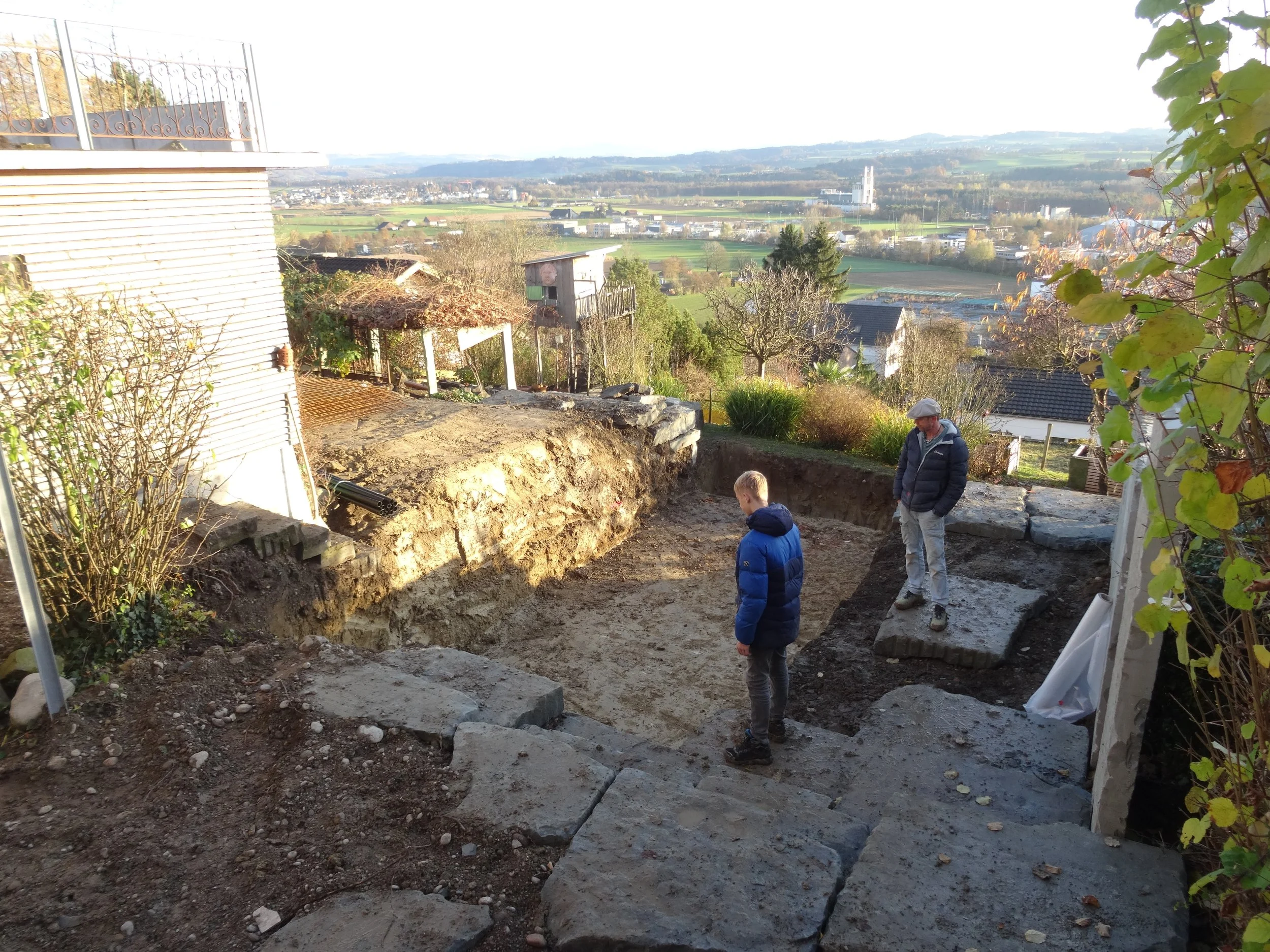 Eine Baustelle im Garten mit einem tiefen Graben, drei Menschen, die das Gelände erkunden, und einer ländlichen Landschaft im Hintergrund.