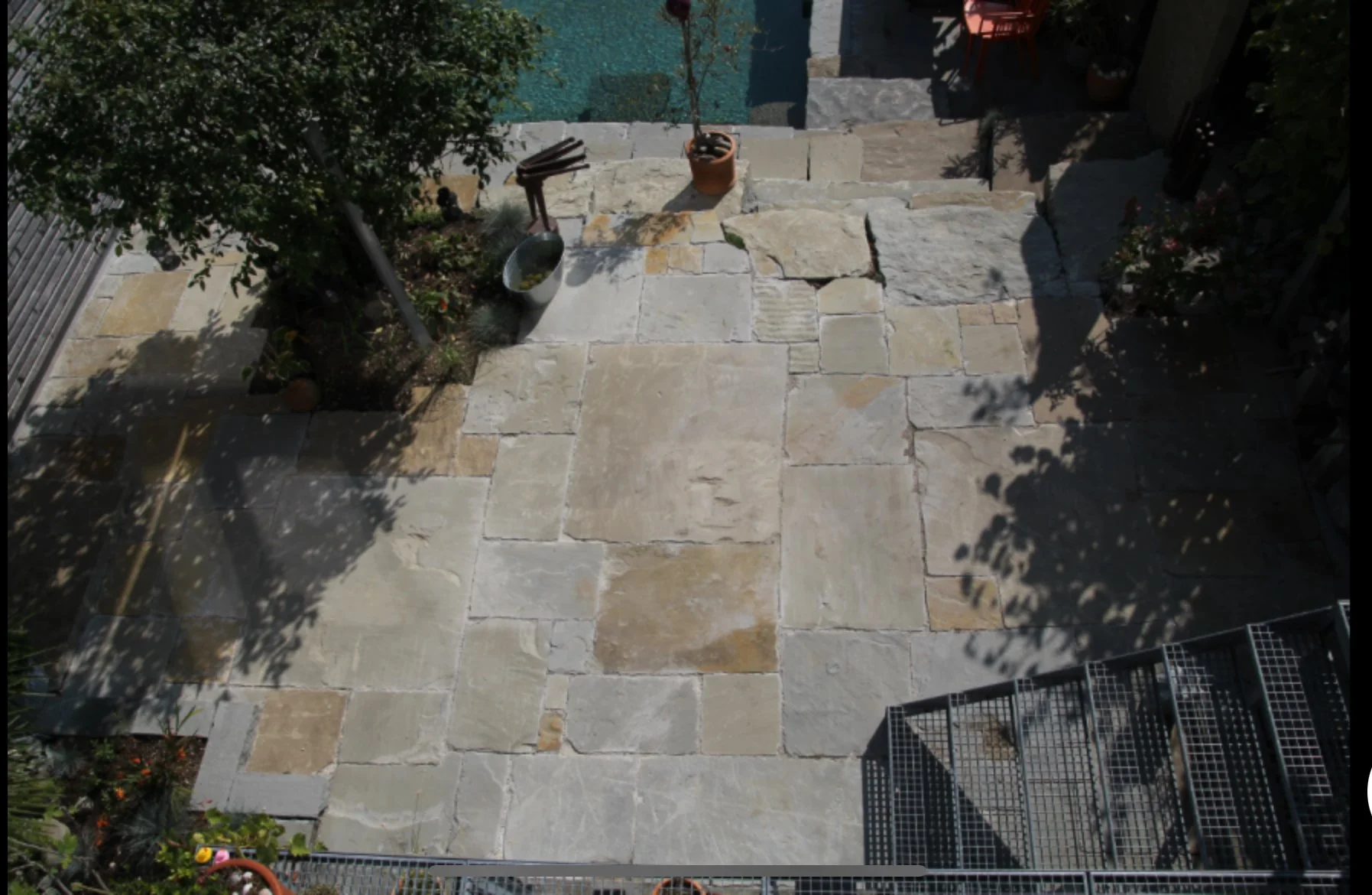 Ein Terrassenboden aus Sandstein Platten, umgeben von Gartenpflanzen und Schatten, mit einem Blumentopf, einer Gartenschaufel und einer kleinen Trittleiter.