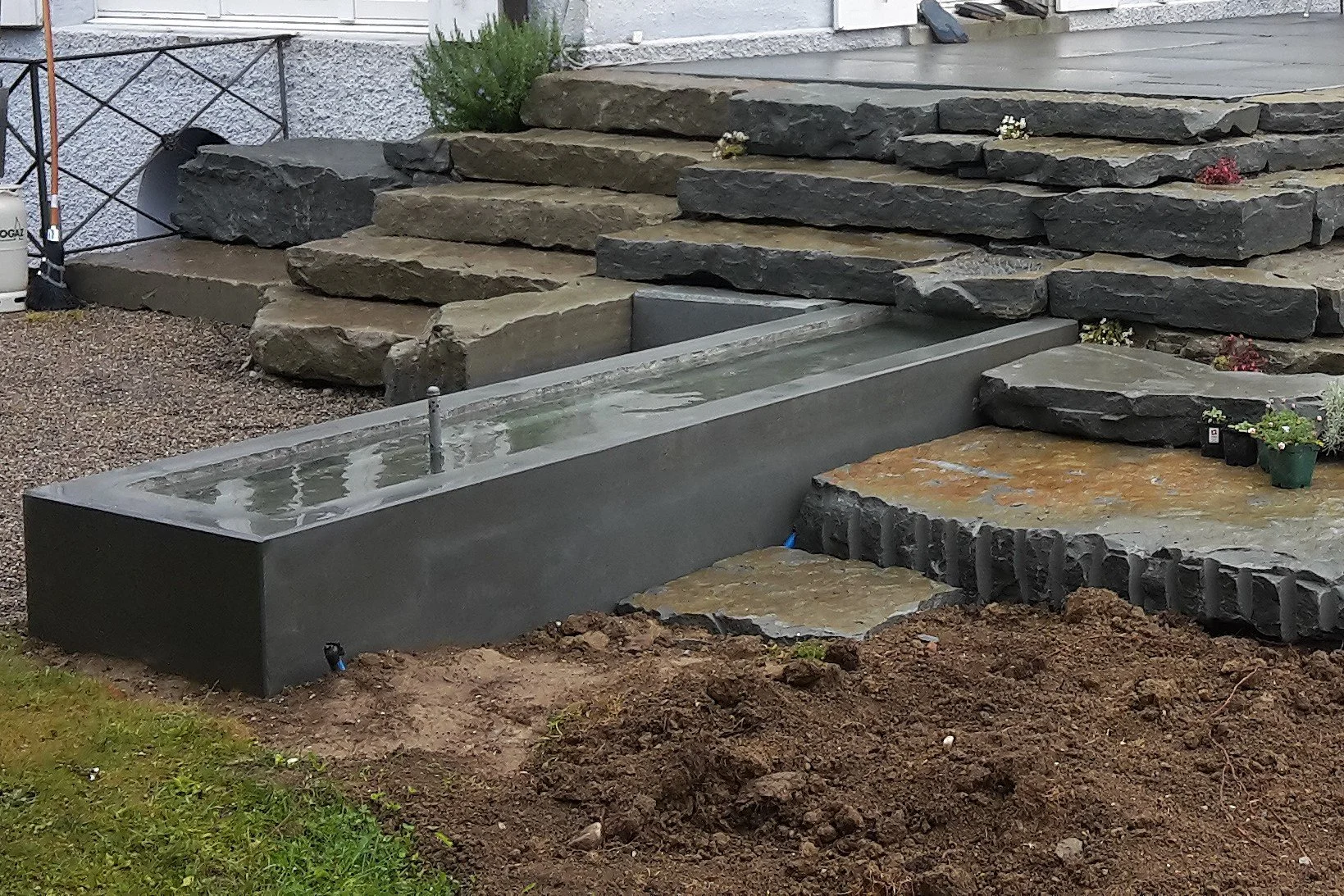 Ein Garten mit einer Wasserquelle in einem rechteckigen, schwarzen Becken, umgeben von unebenen, großen Natursteinplatten, die eine Treppe bilden, und ein Bereich mit Erde und Pflanzen