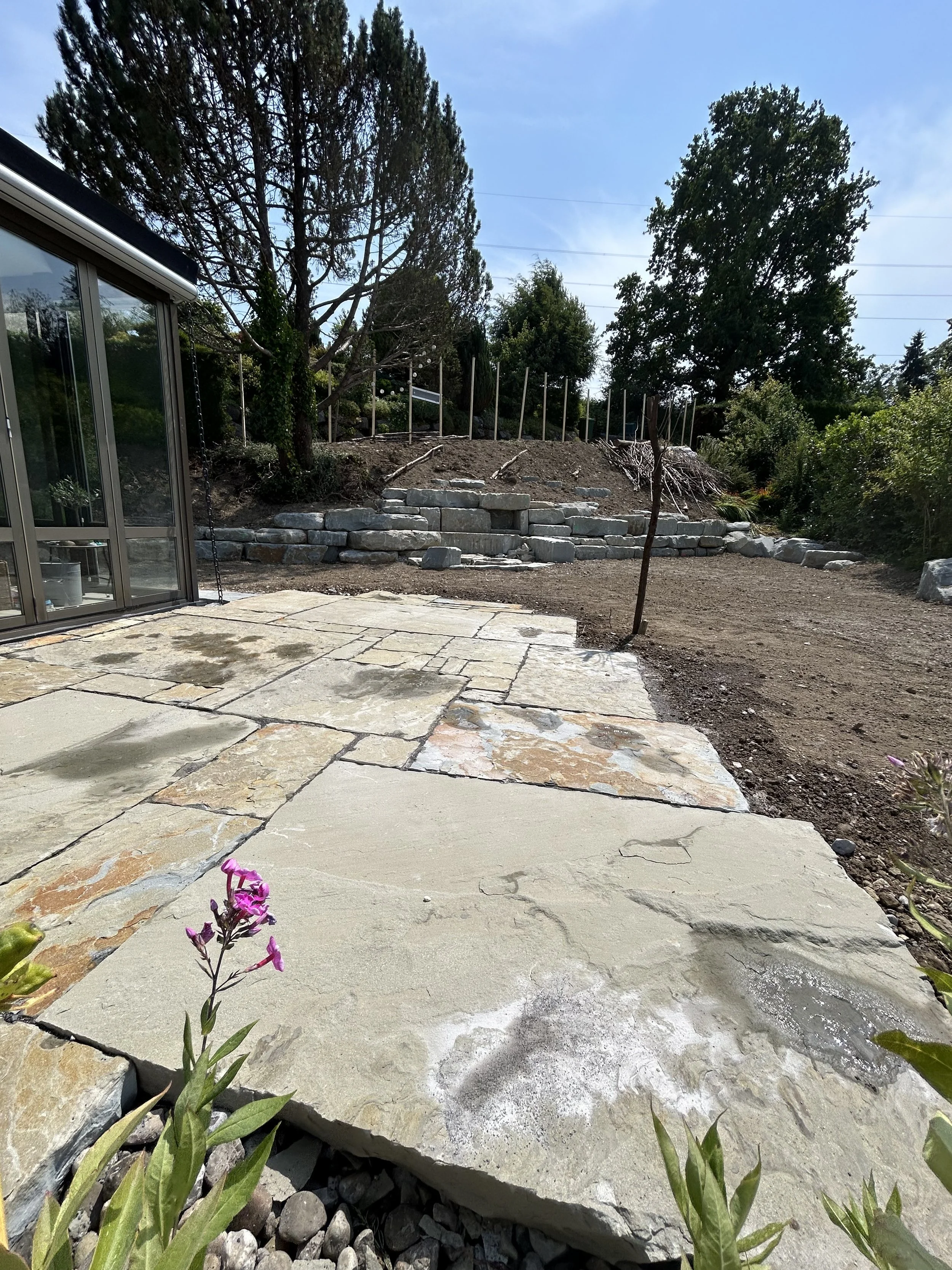 Garten mit Sandsteinplatten, einige Pflanzen im Vordergrund, im Hintergrund ein Garten mit Stufen aus Steinblöcken, Bäumen und einer hohen Hecke.