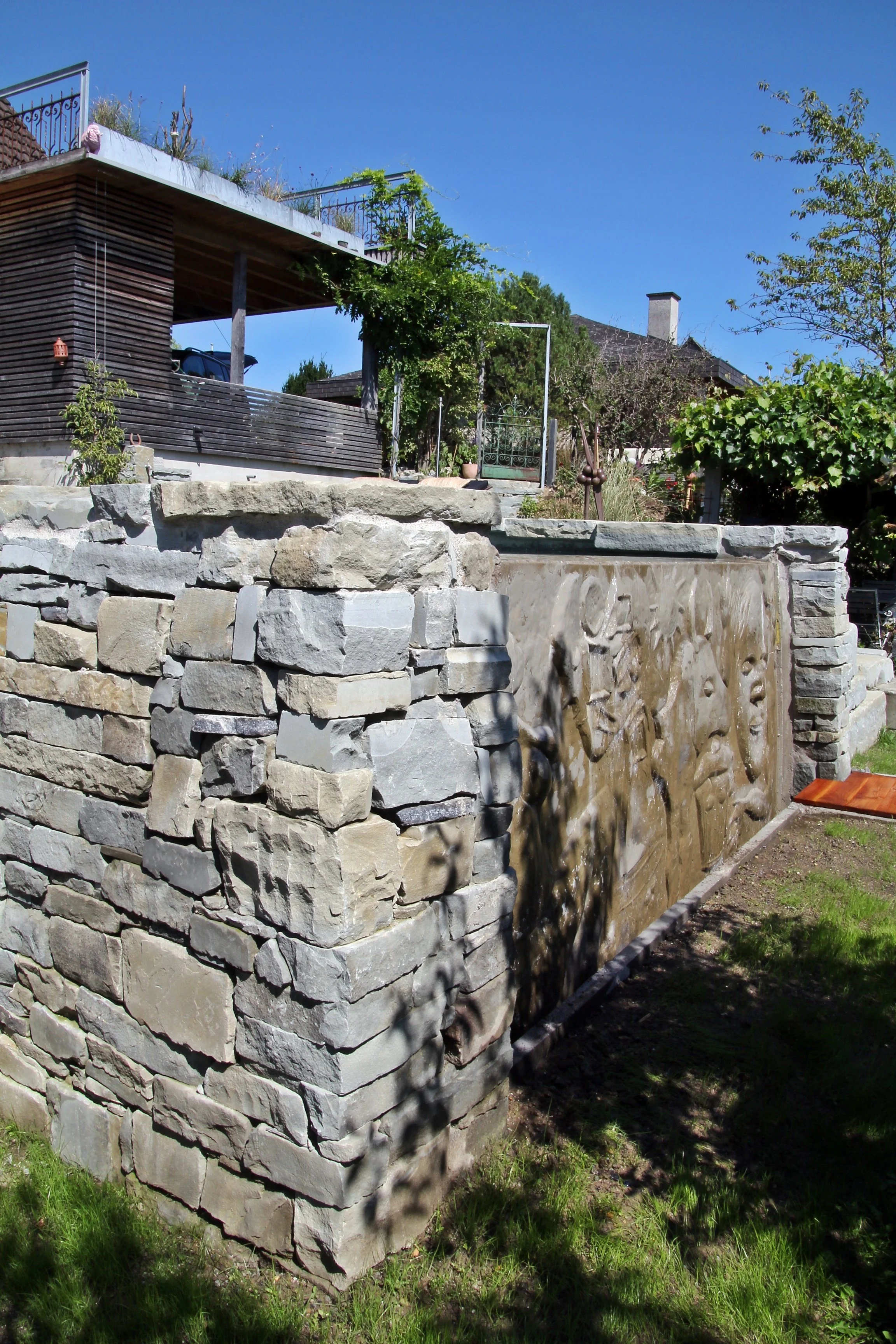 Ein Garten mit einem Wasserfall, der in eine Steinmauer fließt, und einer Terrasse im oberen Bereich mit Gartenpflanzen und einem blauen Himmel.