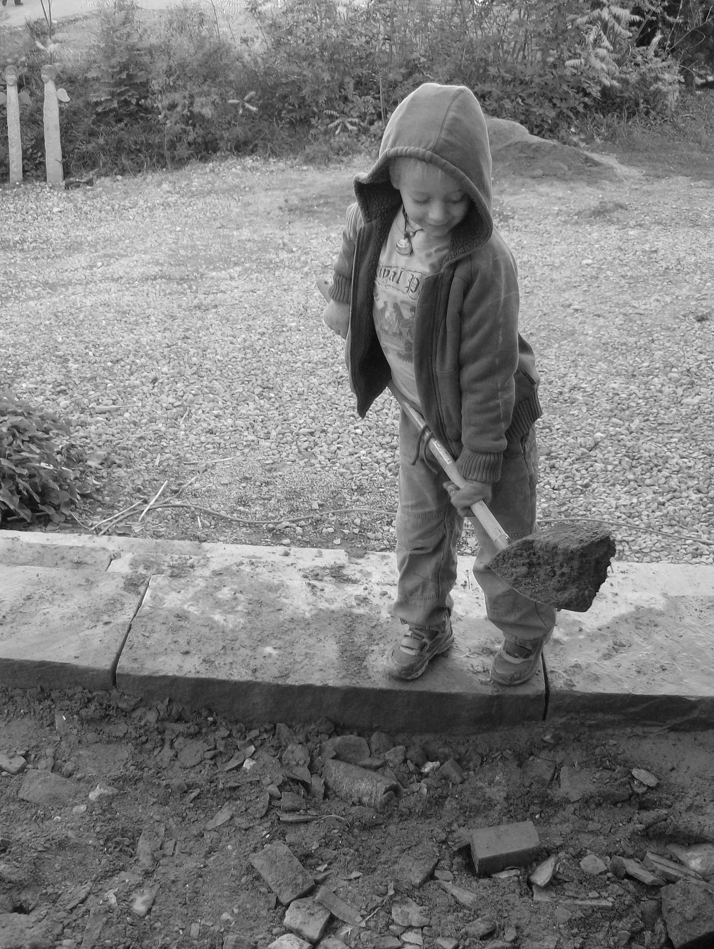 Ein Junge trägt einen Spaten und arbeitet im Garten auf einer Baustelle, umgeben von Erde und Steinen.