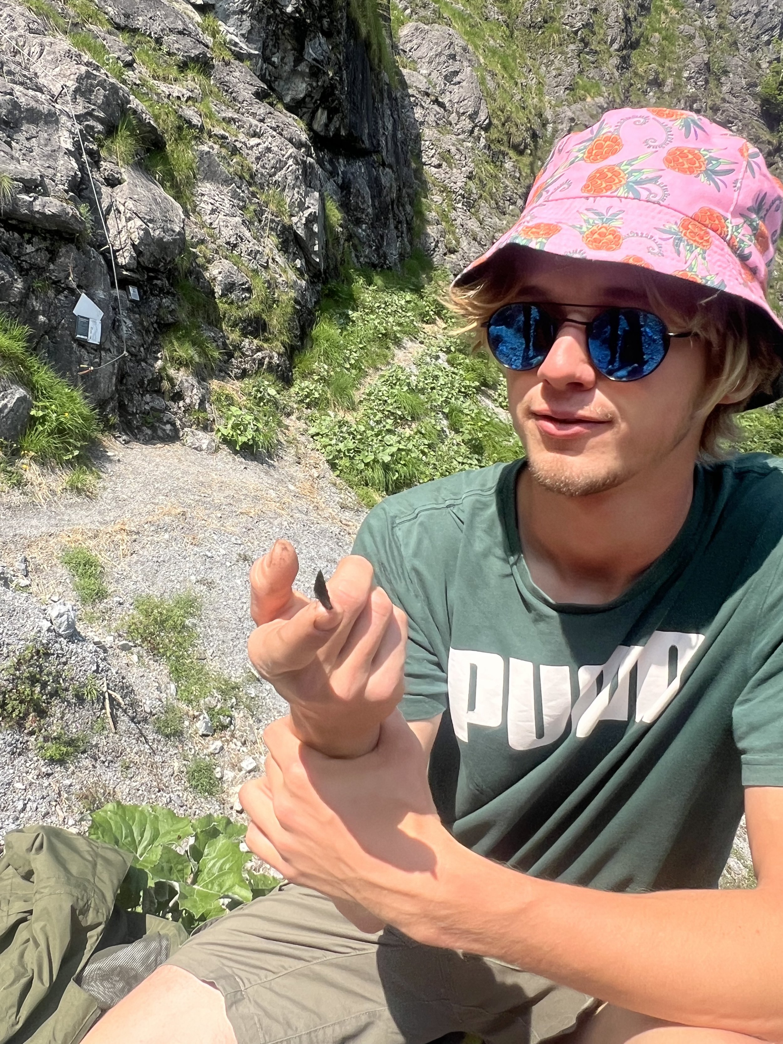 Ein Mann trägt einen pinkfarbenen Hut mit Ananas-Muster, dunkle Sonnenbrille und ein grünes T-Shirt, sitzt im Freien vor einer Felswand, hält einen kleinen Gegenstand in der Hand.