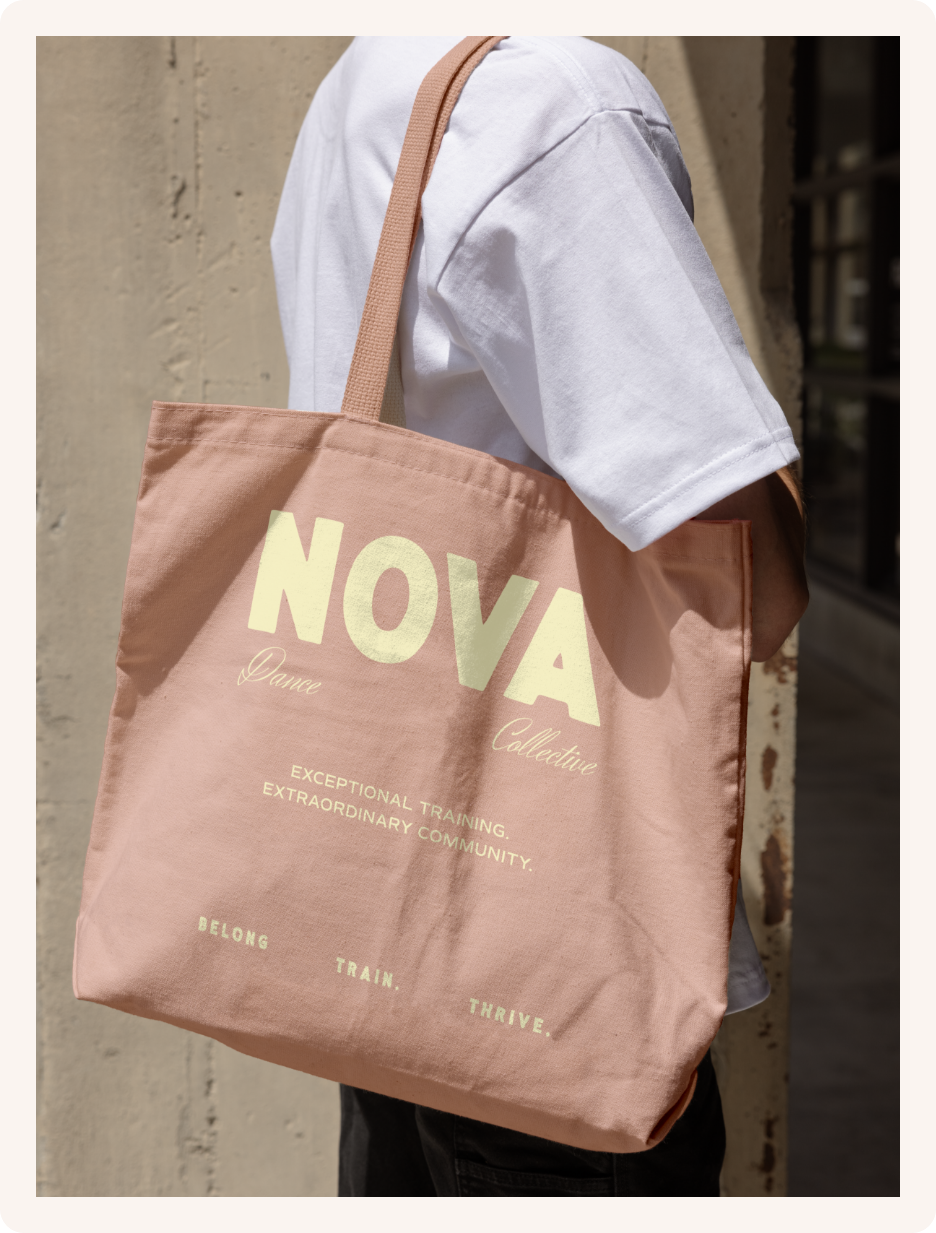 Person wearing a white t-shirt carrying a large pink tote bag with the words 'NOVA Dance Collective' printed on it, standing against a beige wall.