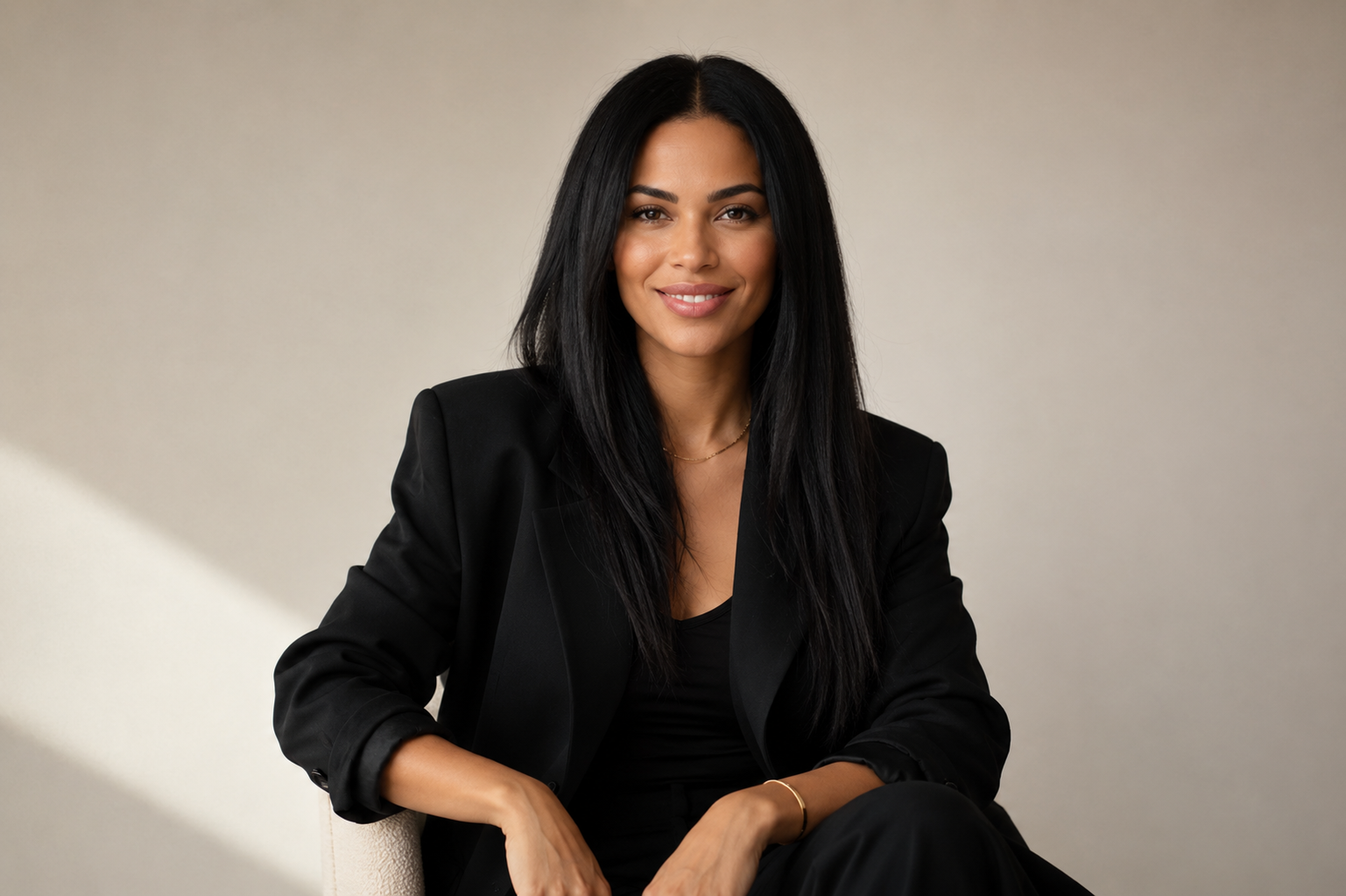 A woman with long black hair wearing a black blazer and black top, sitting and smiling at the camera against a beige background.
