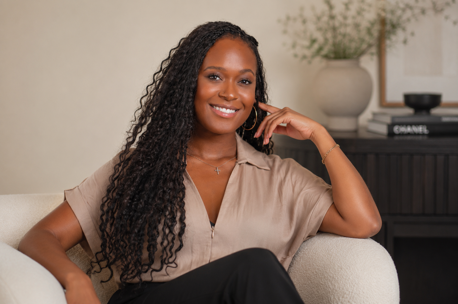 A woman sitting on a white couch, smiling, with a beige blouse, curly hair, and jewelry, in a room with decor including a large vase with greenery and books.