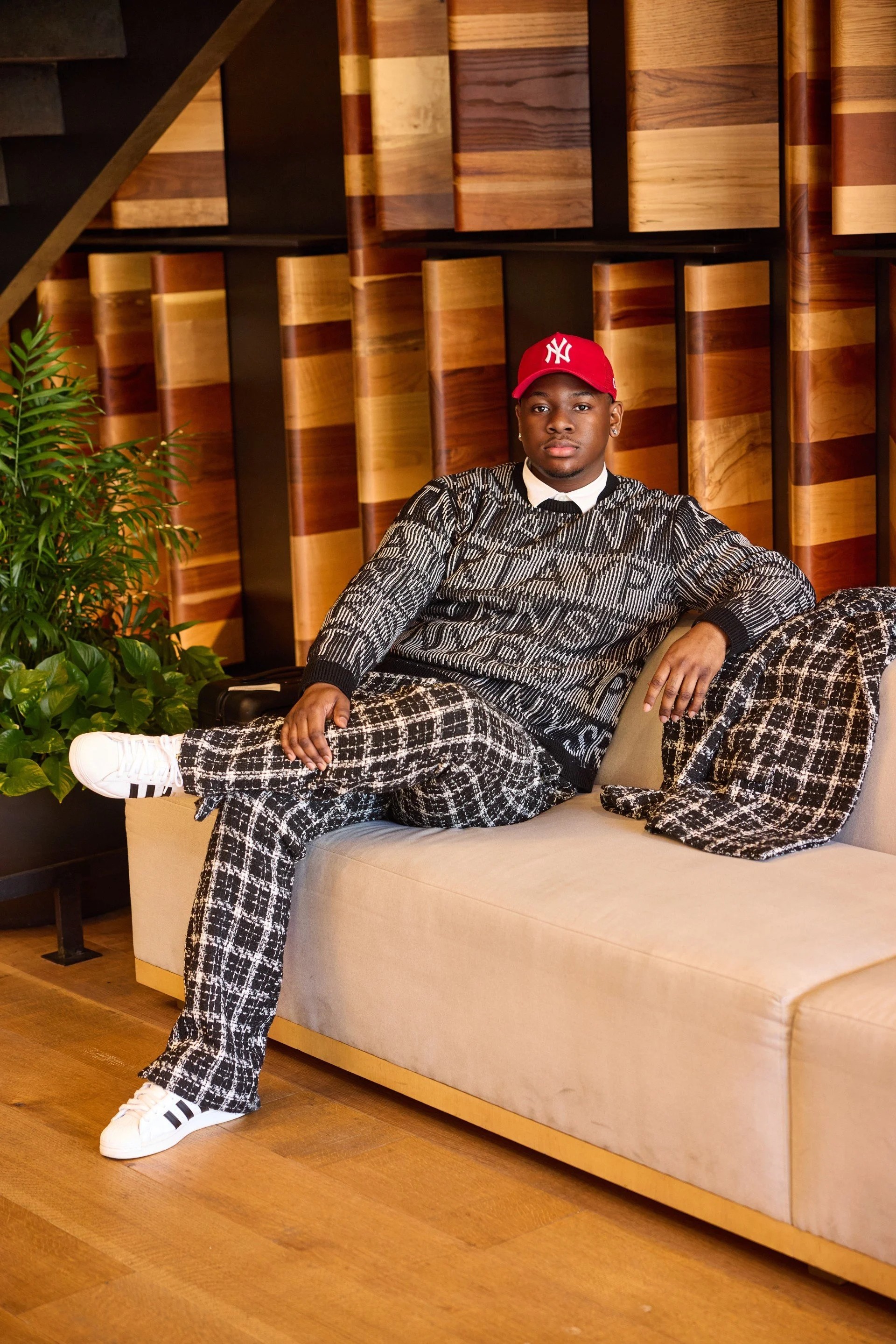 Young man sitting on a beige sofa in a modern room with wooden wall panels, a green potted plant, and wearing a red New York Yankees cap, patterned sweater and plaid pants.