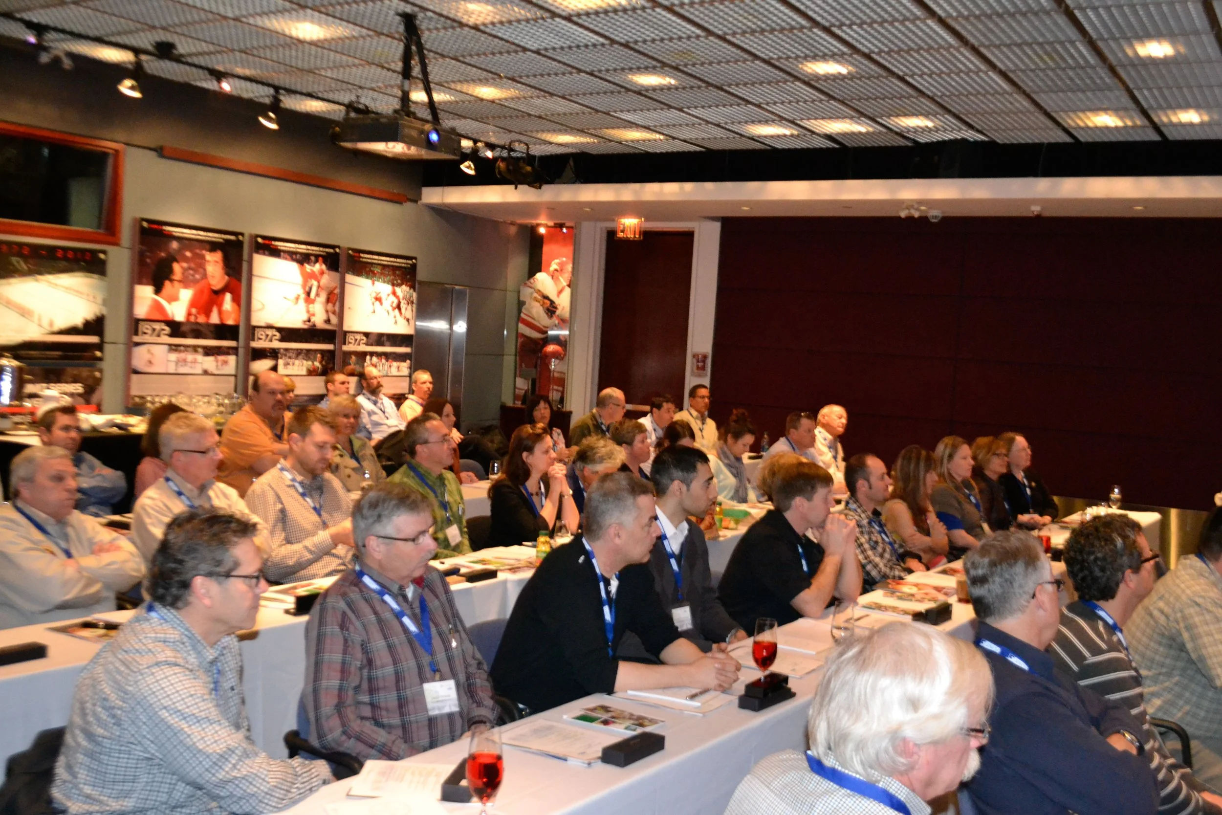 A large group of people seated at tables in a conference room, attentively listening to a presentation or speaker. The room has a high ceiling with a grid pattern and decorated walls with posters or photographs. Some attendees have drinks and papers 