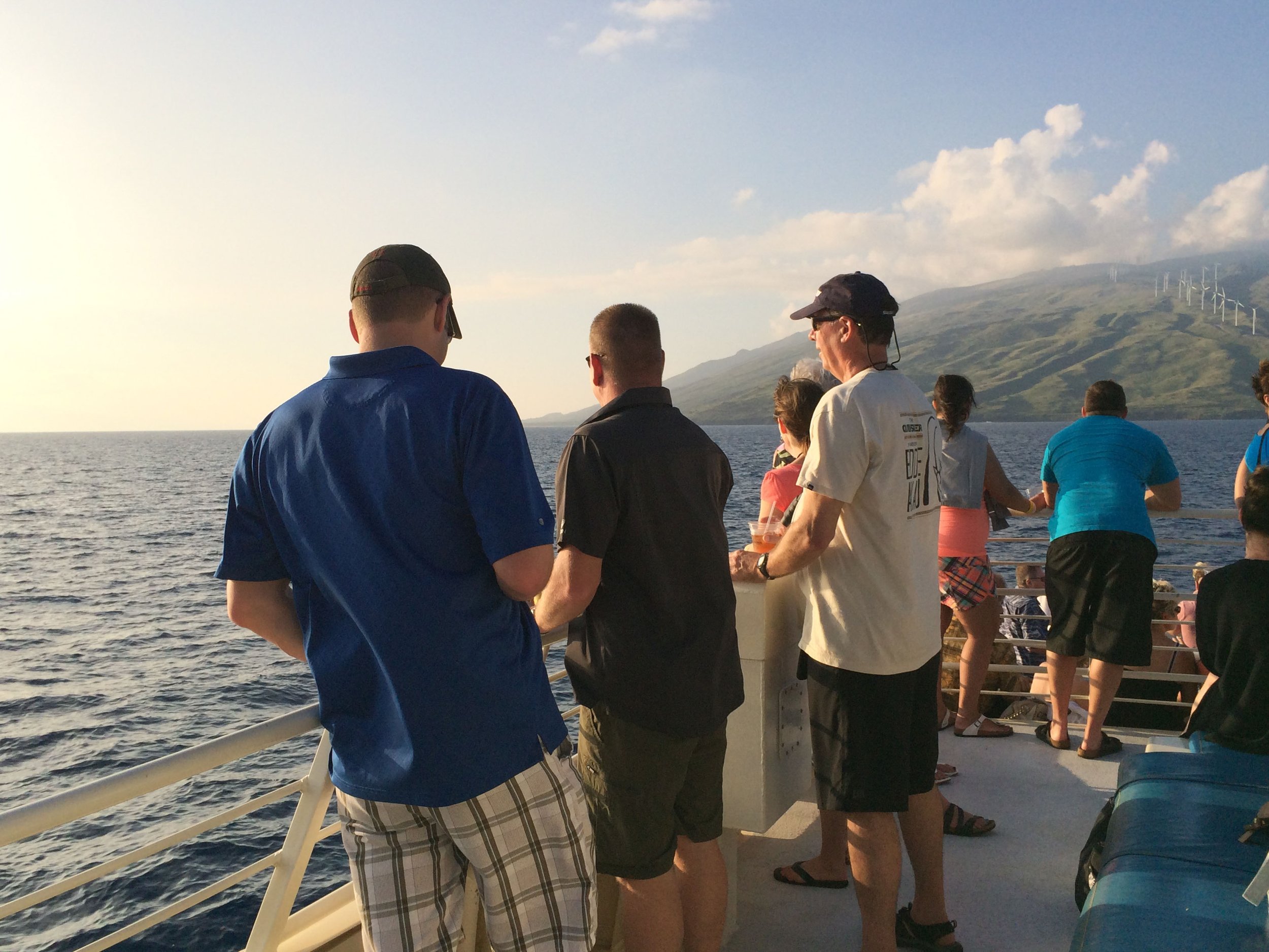 People on a boat looking at the ocean and distant hills with wind turbines under a partly cloudy sky.
