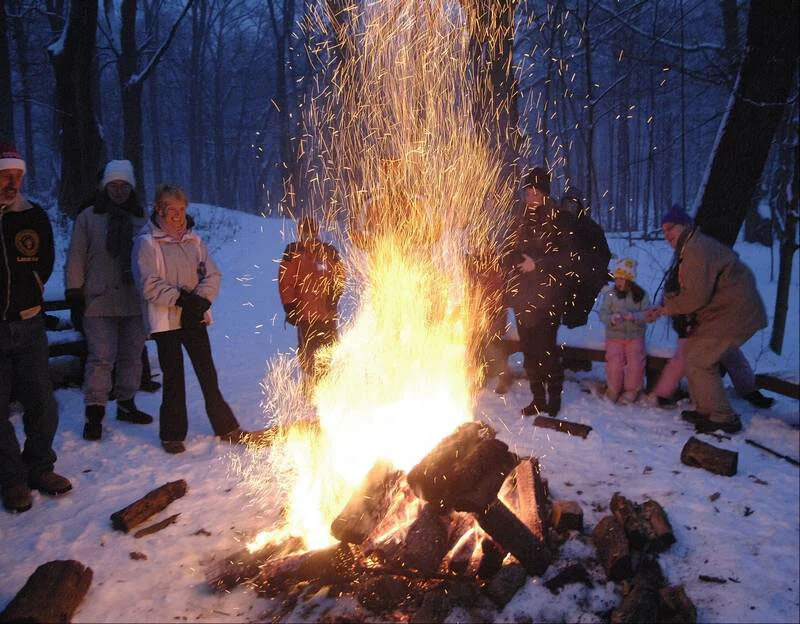 People gathered around a large campfire in a snowy forest, enjoying a winter outdoor gathering.