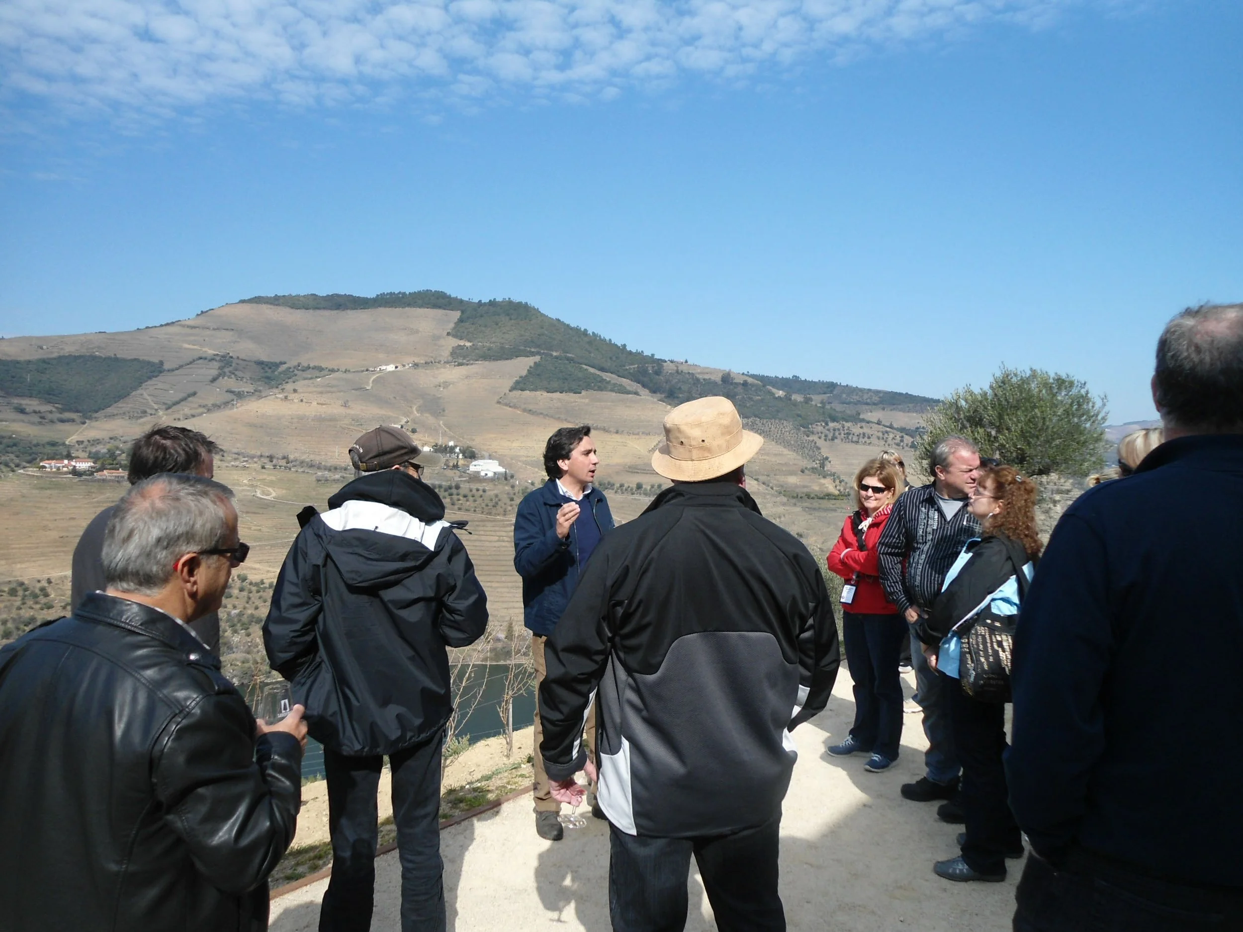 A group of people outdoors with a hill or mountain in the background, some engaged in conversation, some listening, with some holding water bottles, under a partly cloudy sky.