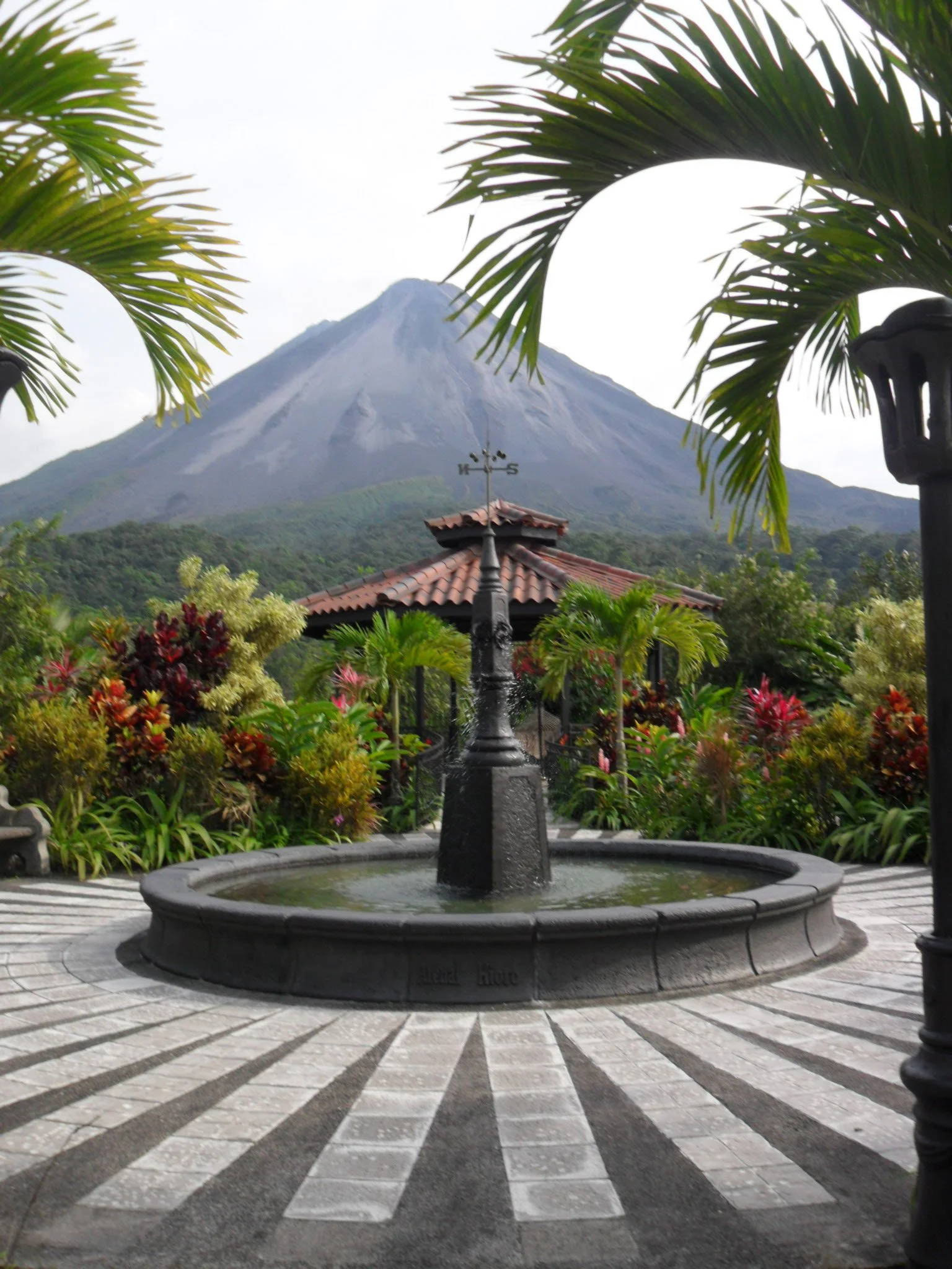 A fountain in a courtyard with a mountain in the background, surrounded by lush greenery and colorful plants.