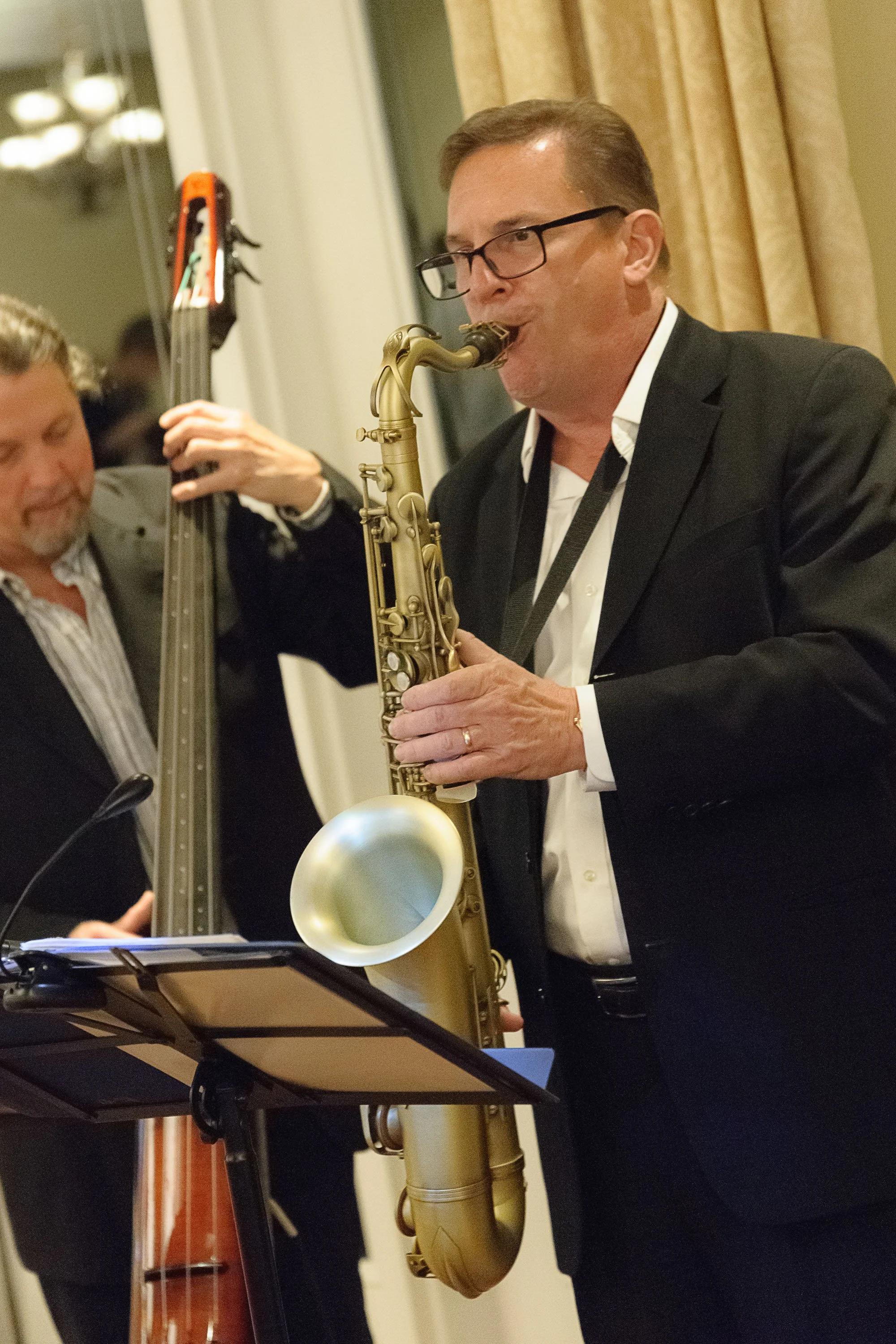 A man wearing glasses in a black suit is playing a saxophone at an indoor event, with a music stand and a person playing a double bass nearby.