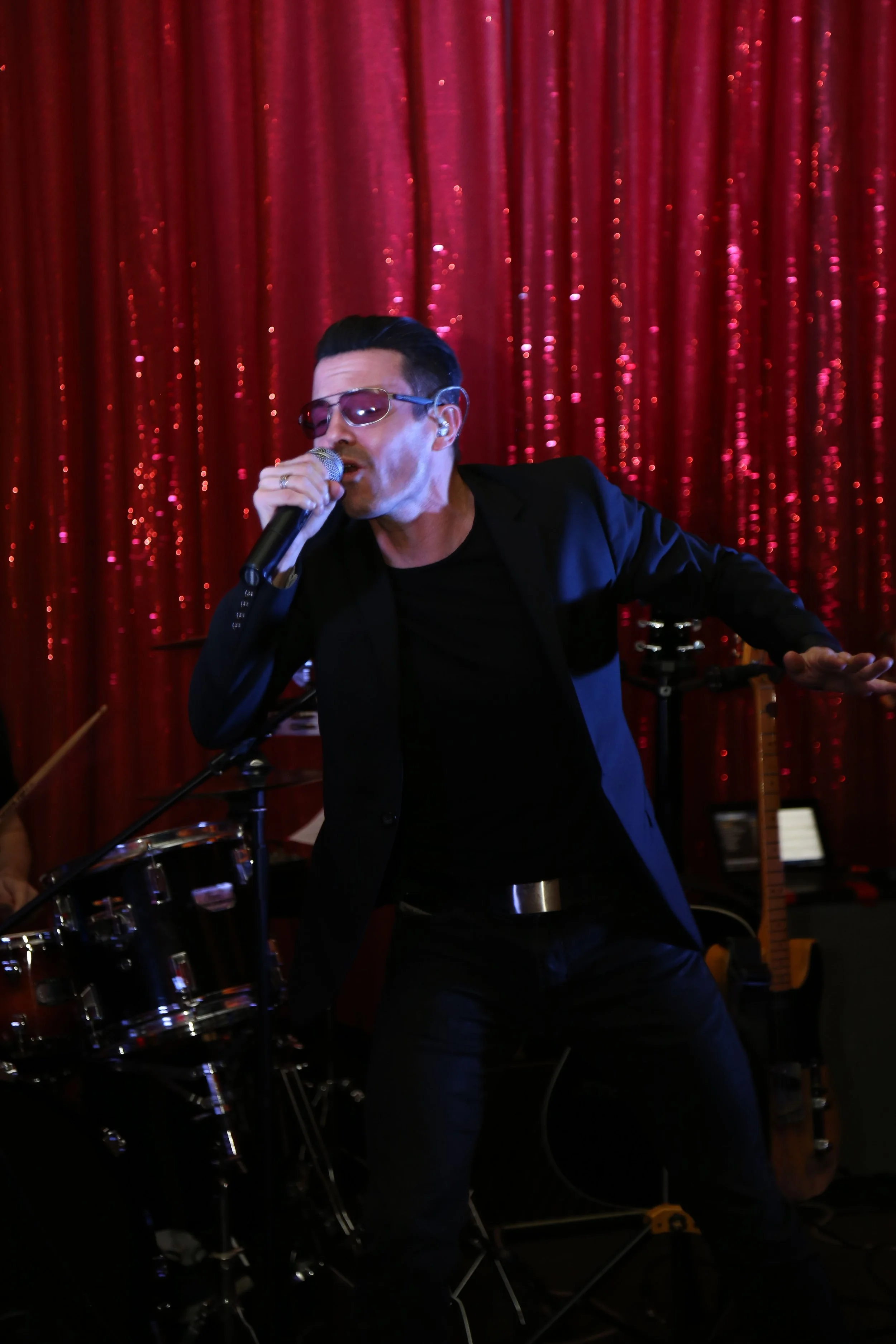 A male singer with dark hair and sunglasses performing with a microphone in front of a red curtain with a shiny, textured appearance, in a music venue with instruments including a drum set and a guitar in the background.