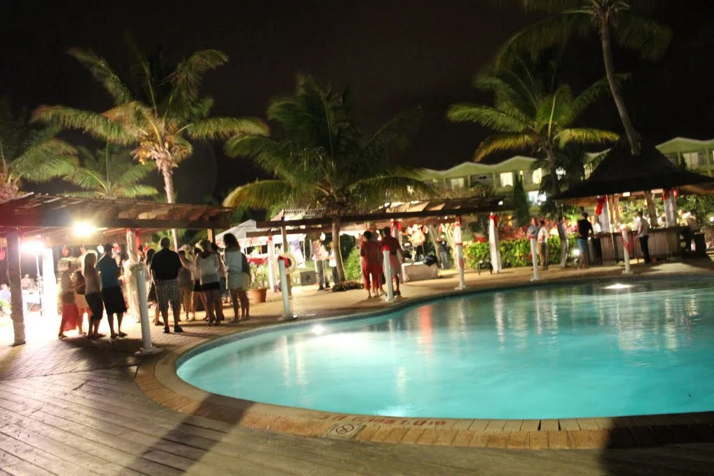 Nighttime scene at a tropical resort with a pool, palm trees, and groups of people gathered under shelters near the pool.
