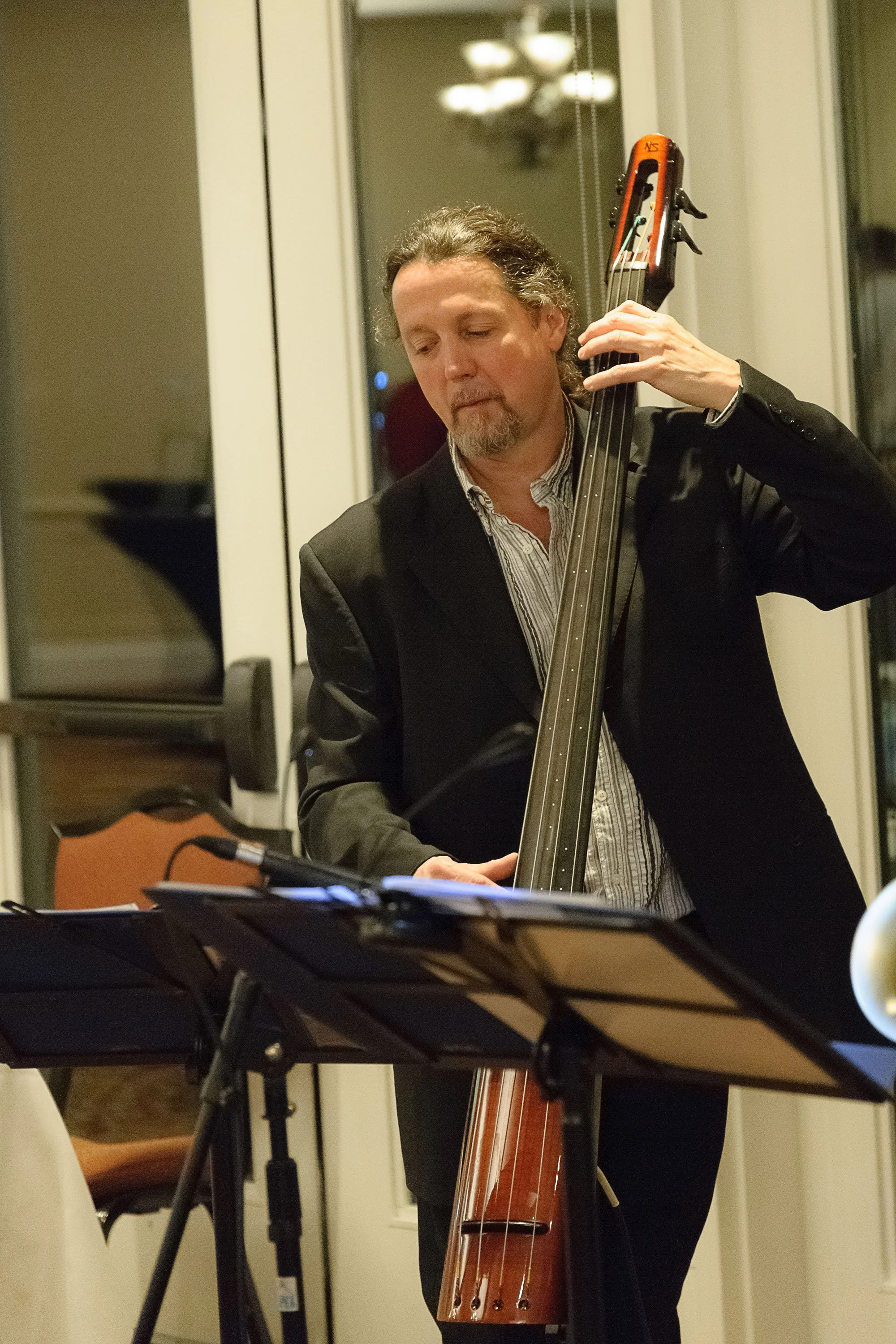 A man with long curly hair and a beard, dressed in a black blazer, playing a double bass in an indoor setting with warm lighting.