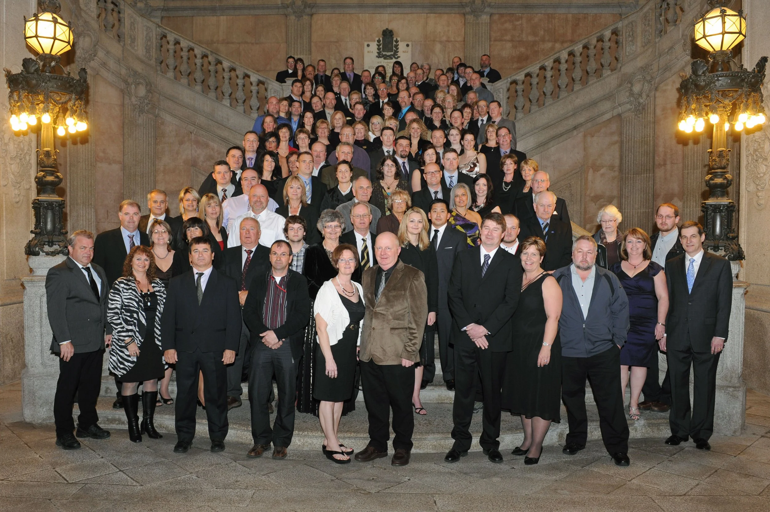 A large group of people dressed in formal attire standing on the steps of a grand staircase in a historic building with ornate lighting and decorative details.