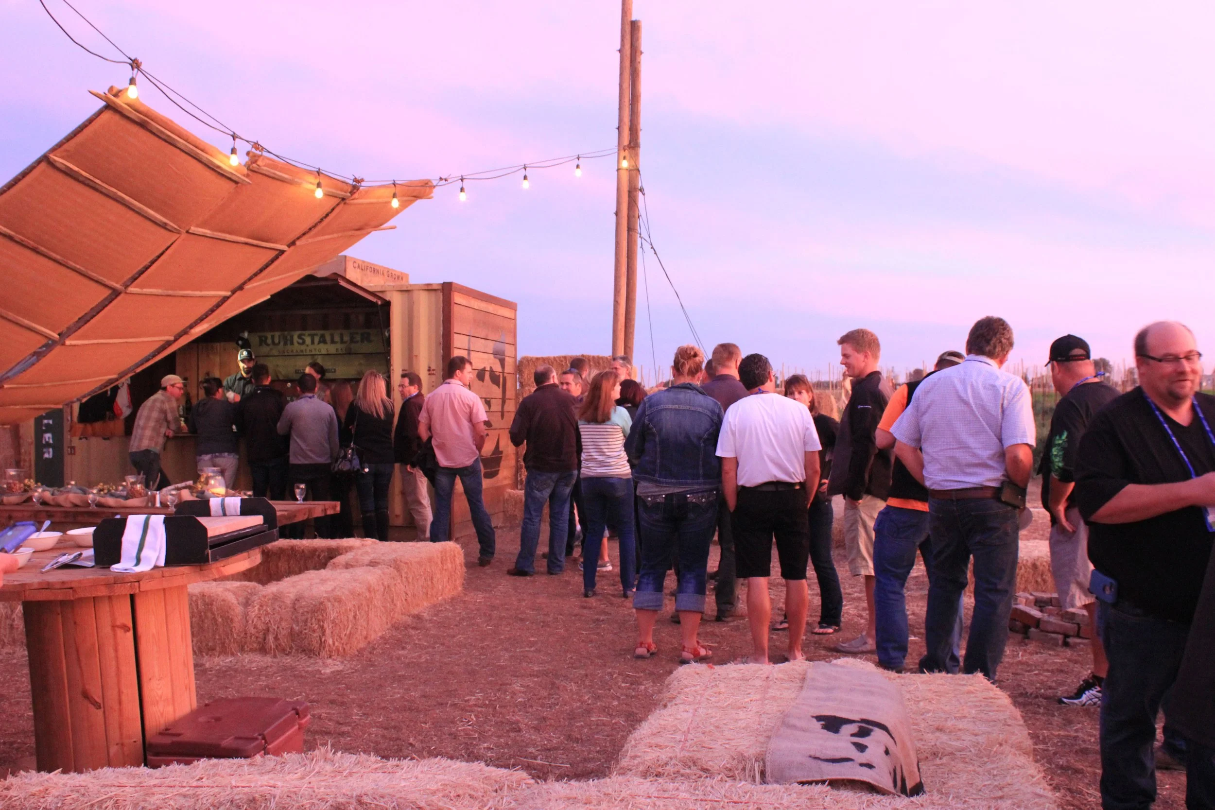 A group of people gathered outdoors during sunset, standing near a food or drink stand with string lights overhead, and straw bales and tables in the foreground.