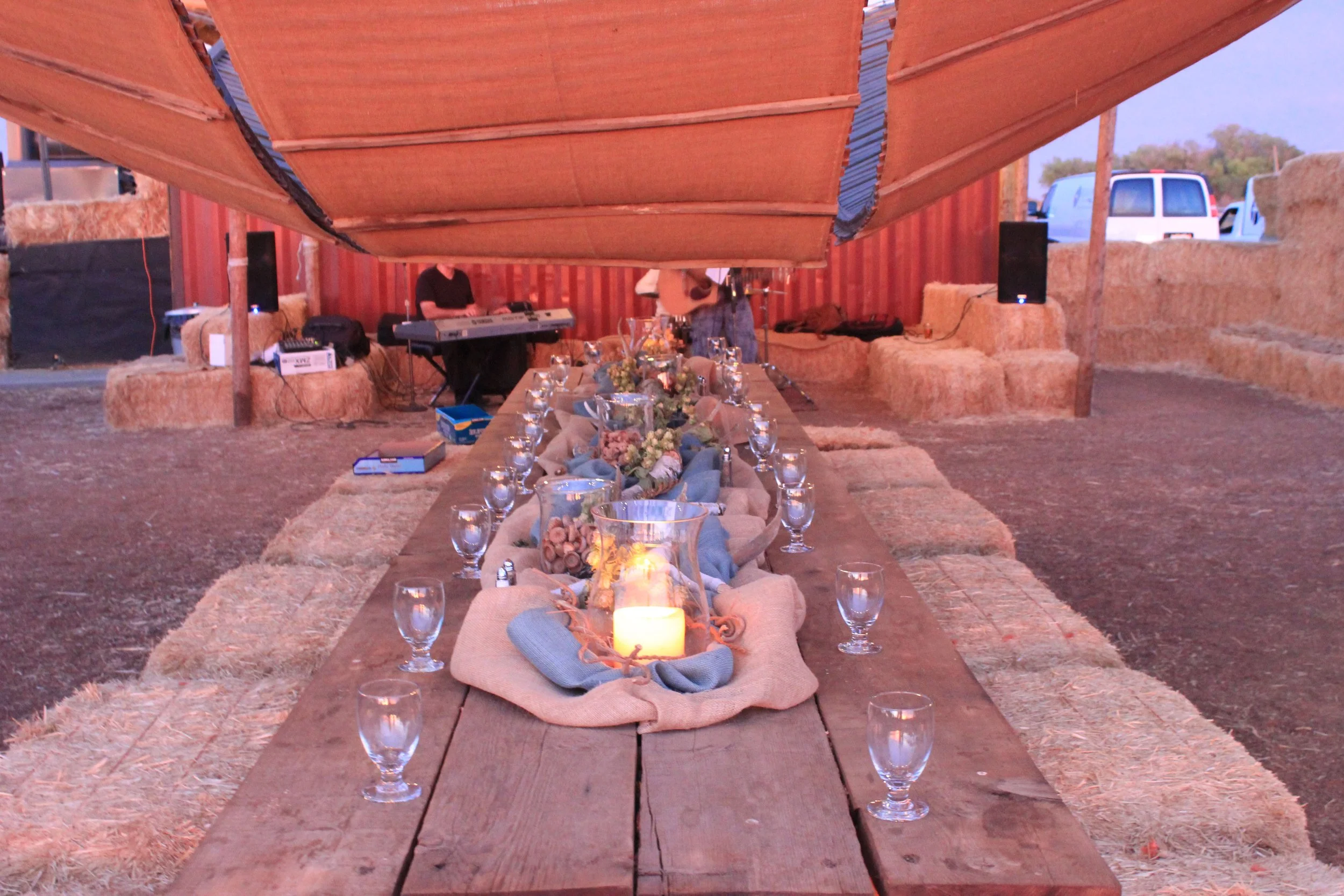 Long wooden table set with glasses, flowers, and candles, surrounded by straw bales, inside a rustic barn with a canopy overhead.