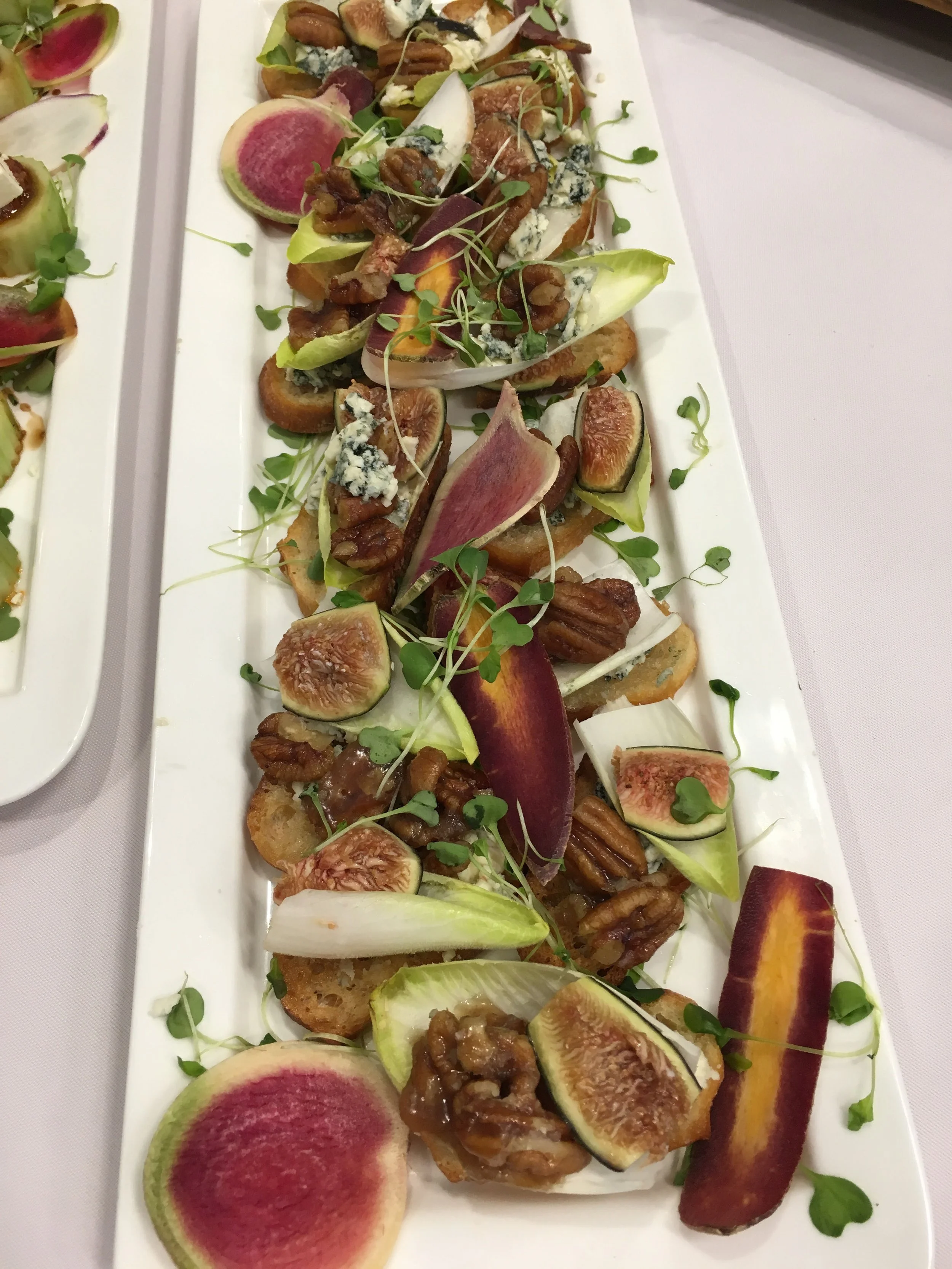 A rectangular white platter featuring slices of fresh figs, watermelon radishes, leafy greens, and small microgreens garnished with nuts and cheese.