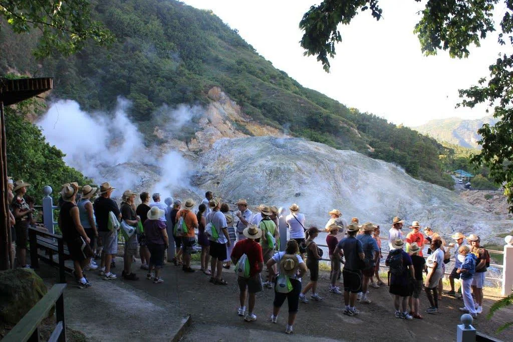 A group of tourists wearing hats and backpacks standing on a viewing platform observing a volcanic hot spring with steam rising from the ground, set against a lush green hillside.