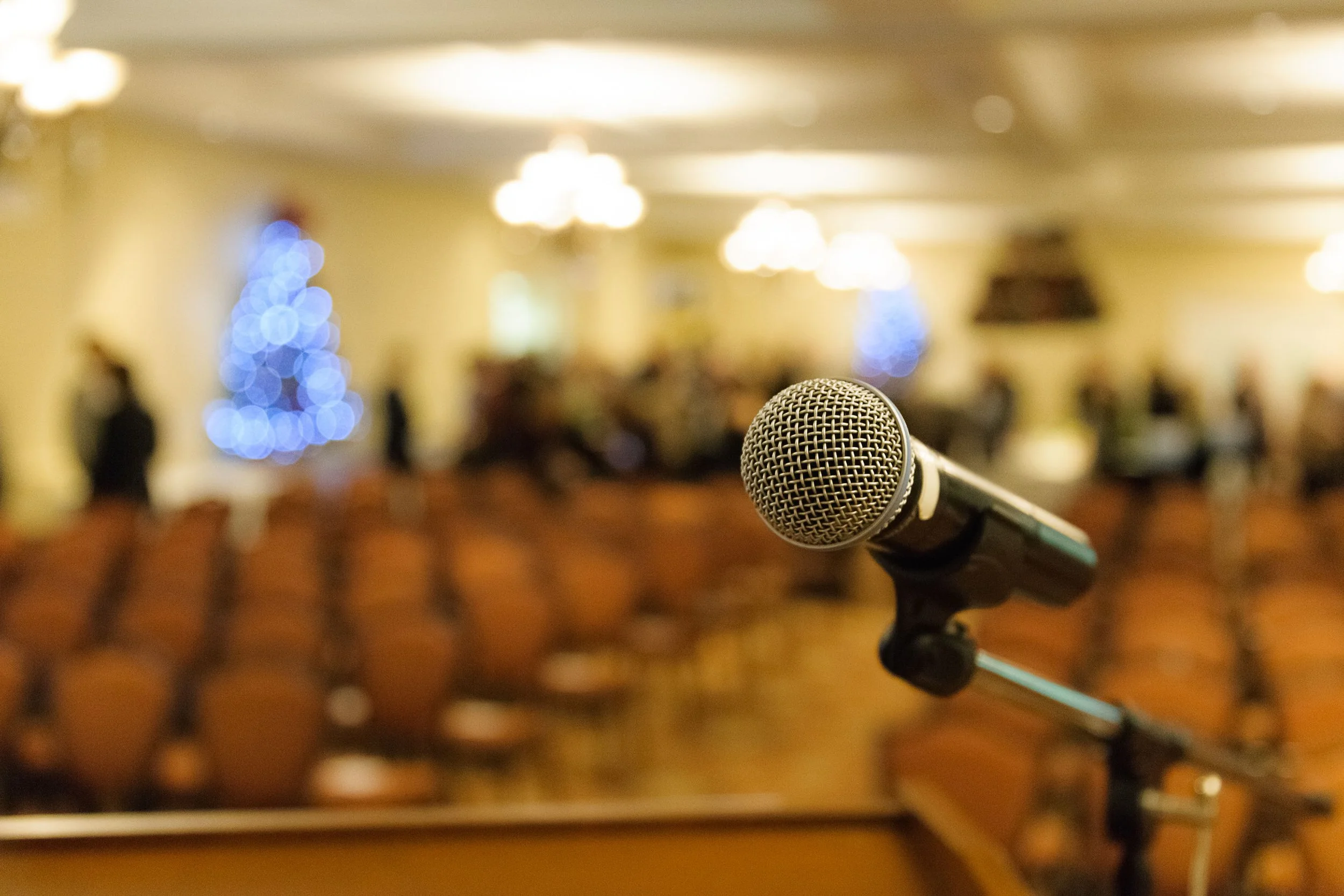 A close-up of a microphone in focus with blurred background of a decorated room, possibly for an event or speech.