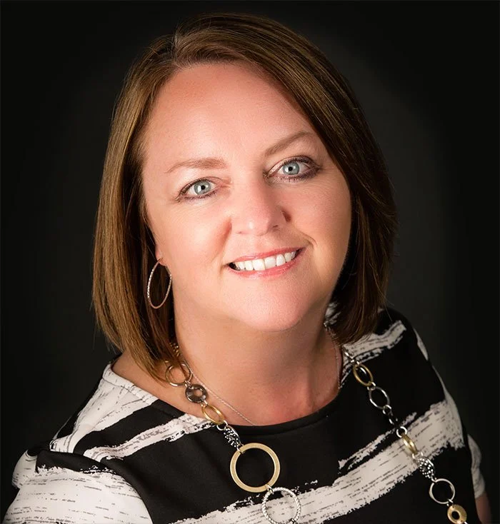 Portrait of a woman with short brown hair, blue eyes, wearing hoop earrings, a black and white striped top, and a decorative chain necklace, smiling against a dark background.
