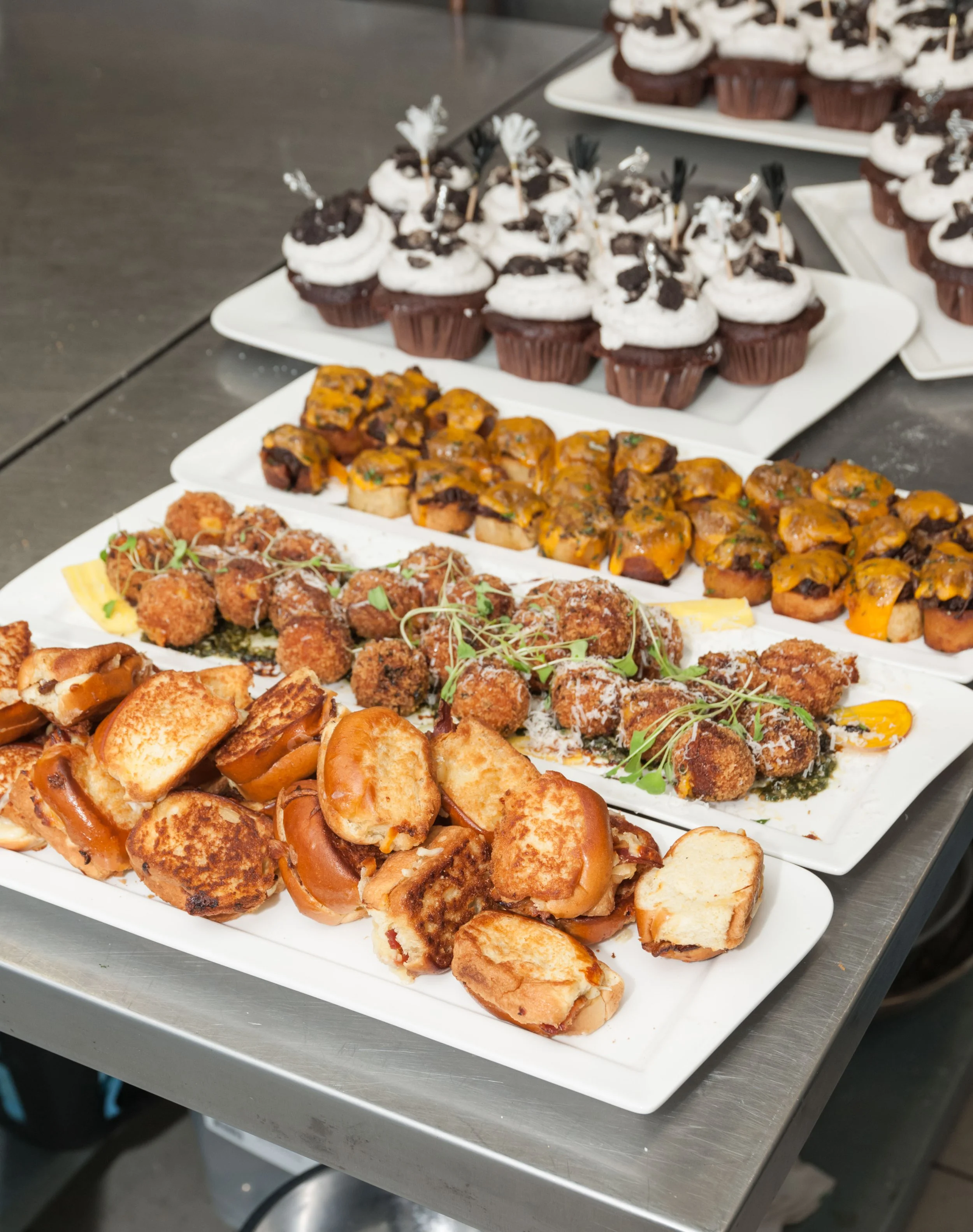 Assorted appetizers, including fried croquettes, mini tacos, and cupcakes with Oreo toppings on white platters.