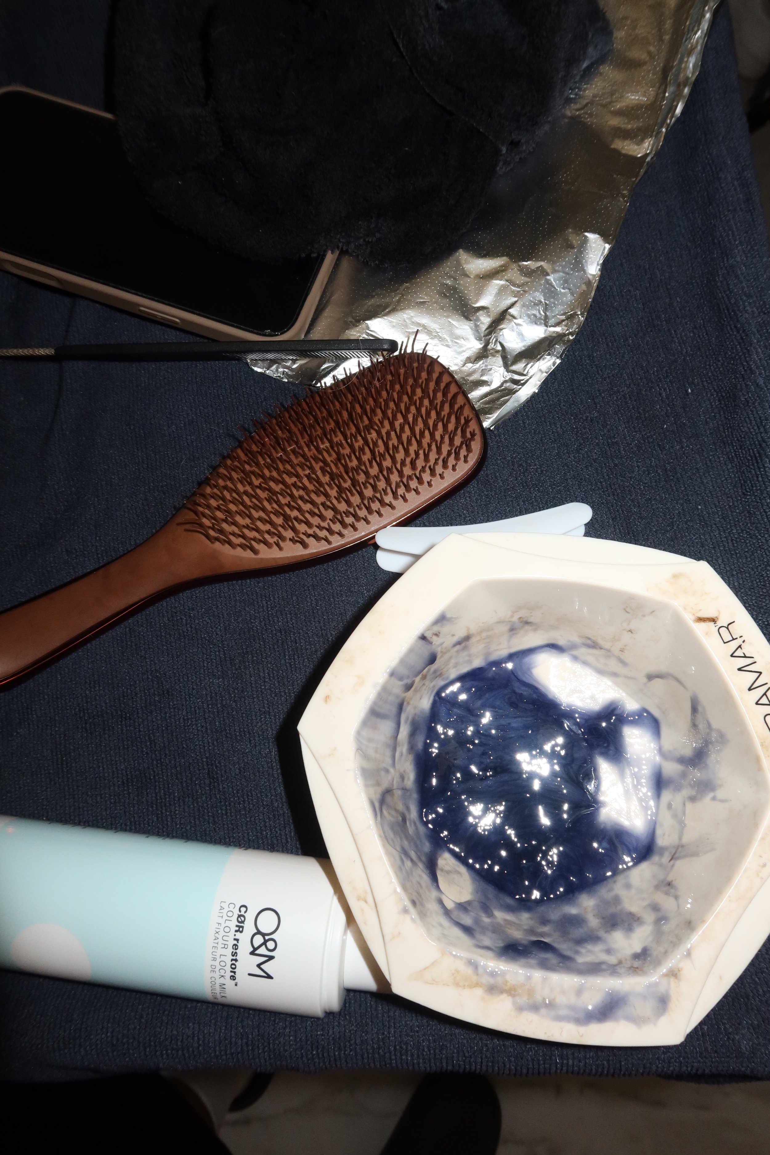 Various personal grooming and skincare items including a bath brush with a wooden handle, a black cloth, a white tube, a black smartphone, a sheet of aluminum foil, a white bowl with a dark blue substance, and some white paper or napkins, all on a dark fabric surface.