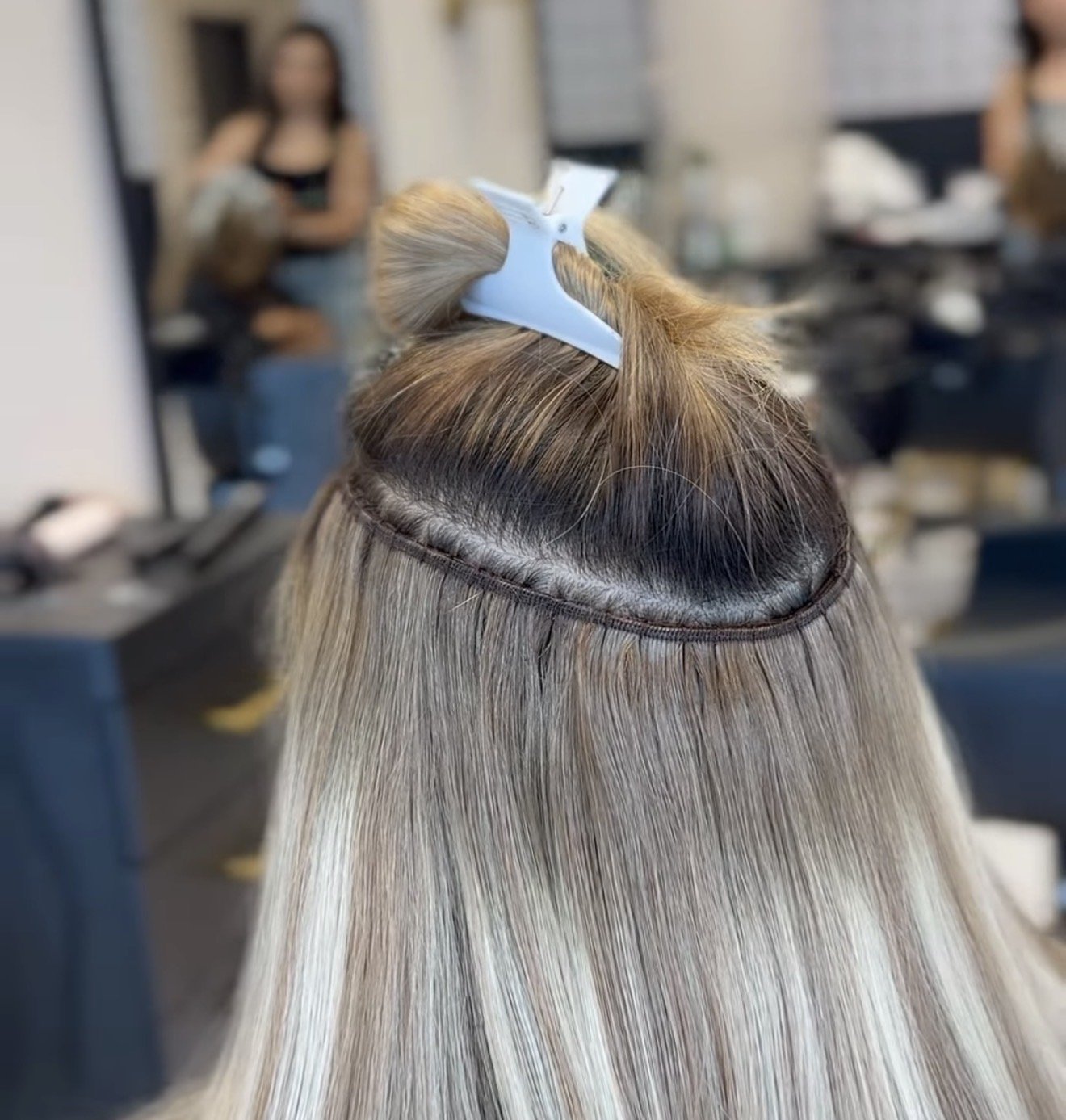 Close-up of a person in a salon with their hair sectioned and secured with a plastic hair clip, while a hairstylist and another person are blurred in the background.