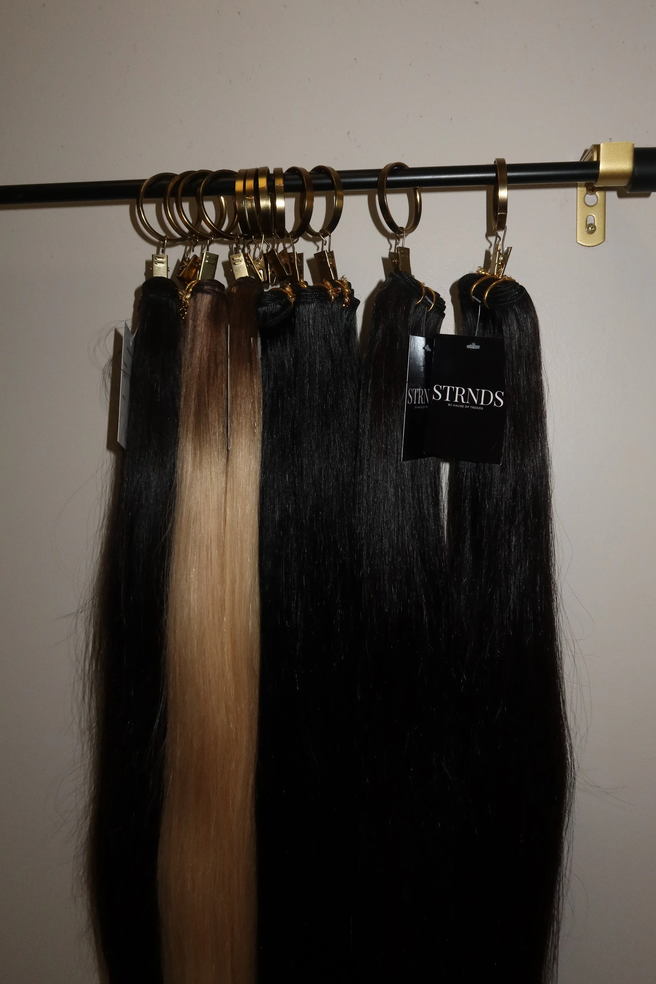 Multiple hair extensions hanging on a black metal rod, each with a gold clip and some with gold tags, in various shades from blonde to dark brown.