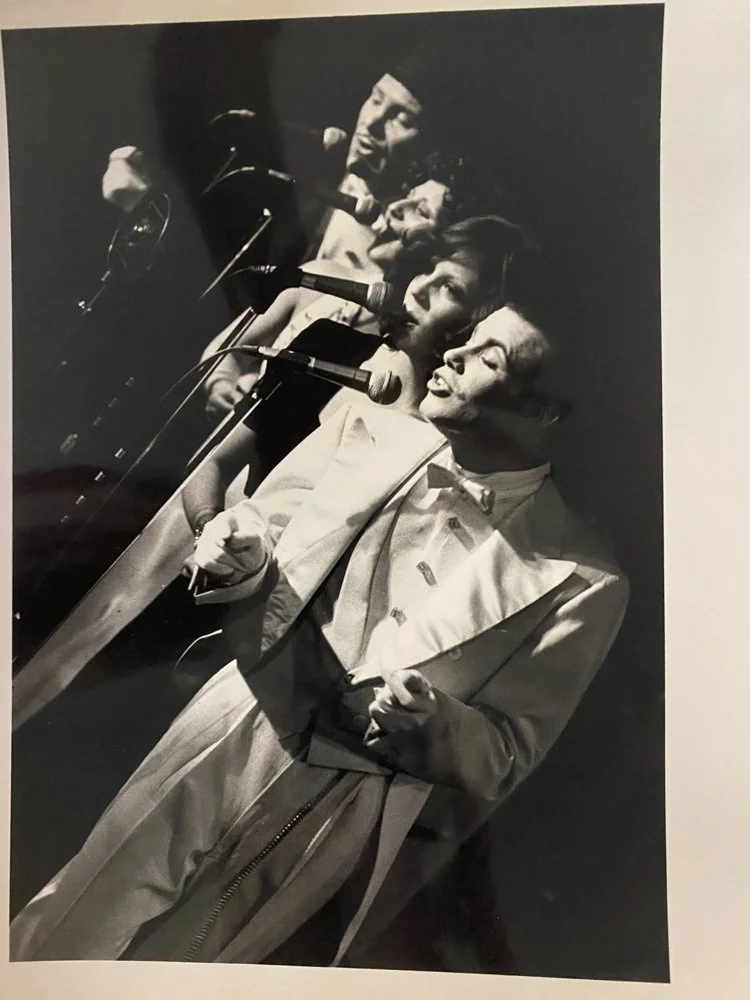Black and white photo of four singers performing on stage with microphones, dressed in vintage clothing, with the lead singer in front wearing a suit with suspenders.