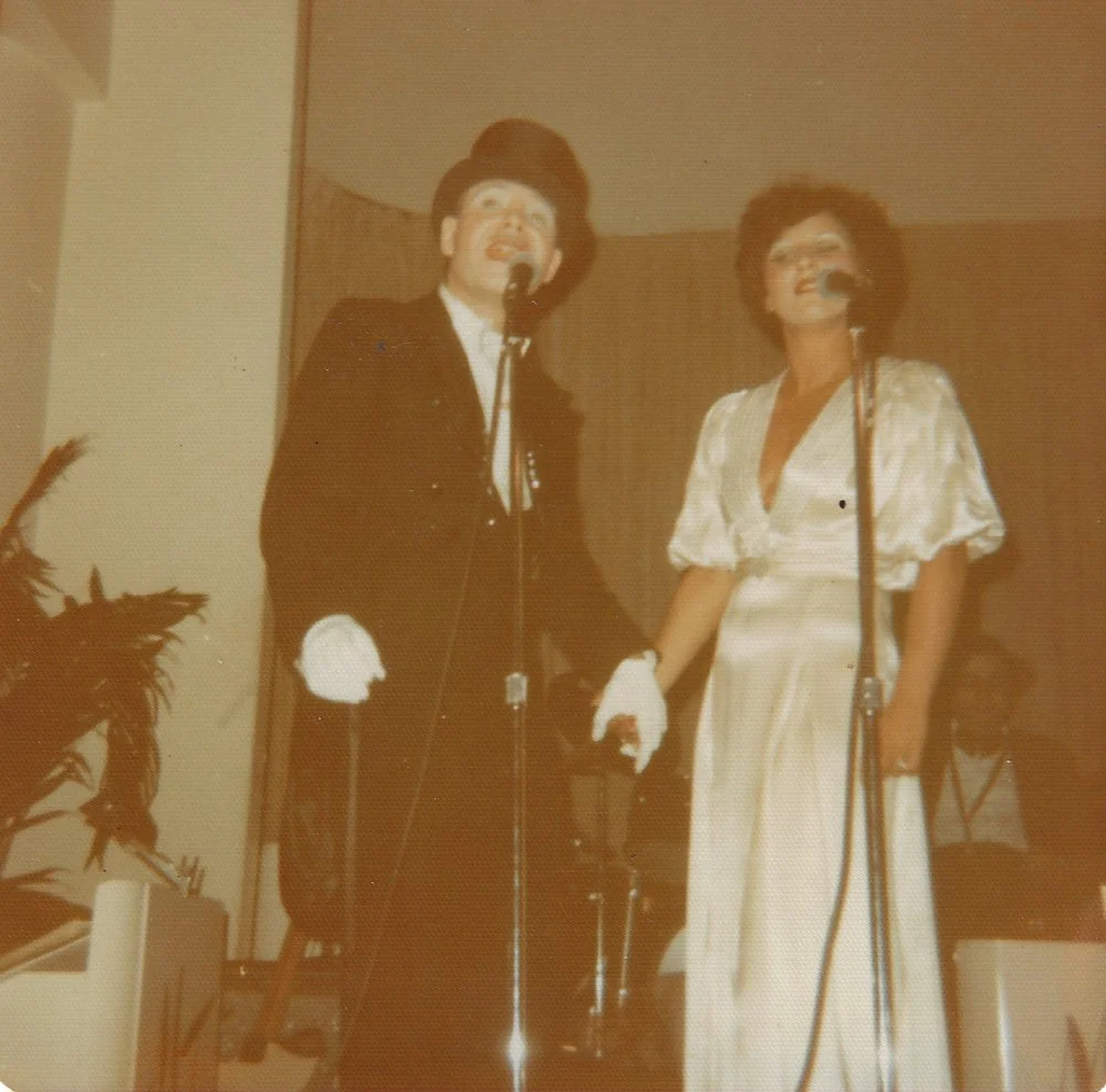 Two people dressed in vintage costumes, holding hands, singing into microphones on a stage.