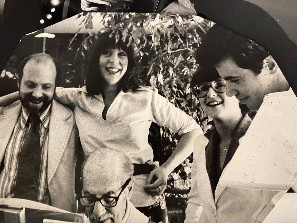 Four people smiling and gathered around a table outdoors, with trees in the background. One person is reading a book, some are wearing glasses, and they appear to be enjoying a social moment.