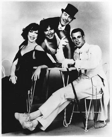 Black and white photo of four people, two women, one man, and one young boy, with the man wearing a top hat. They are smiling and posing with a table, in vintage clothing style.