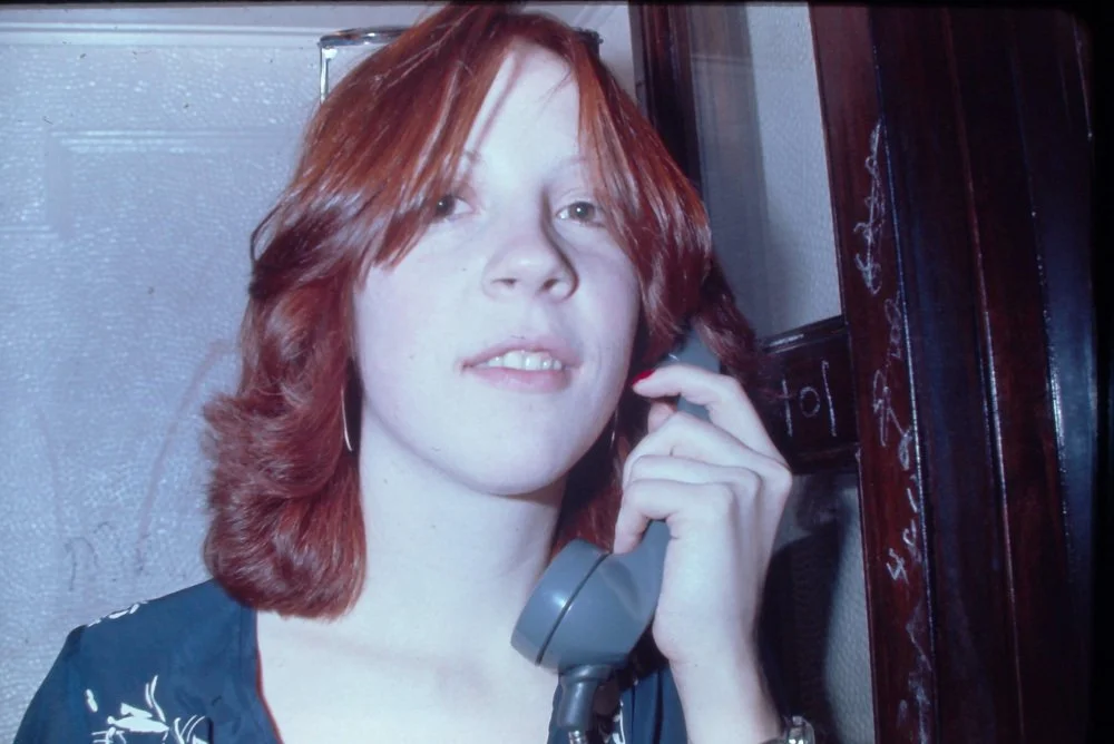 A young woman with shoulder-length red hair talking on a gray landline phone indoors.