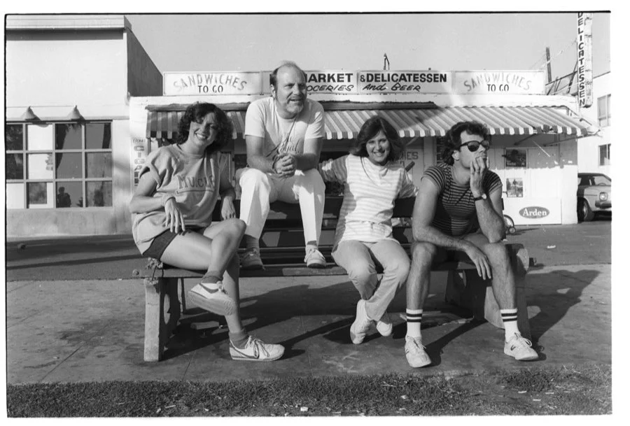 Group of four young friends sitting on a bench outside a store with signs that read 'Sandwiches to go' and 'Market & Delicatessen' in the background, black and white photo.