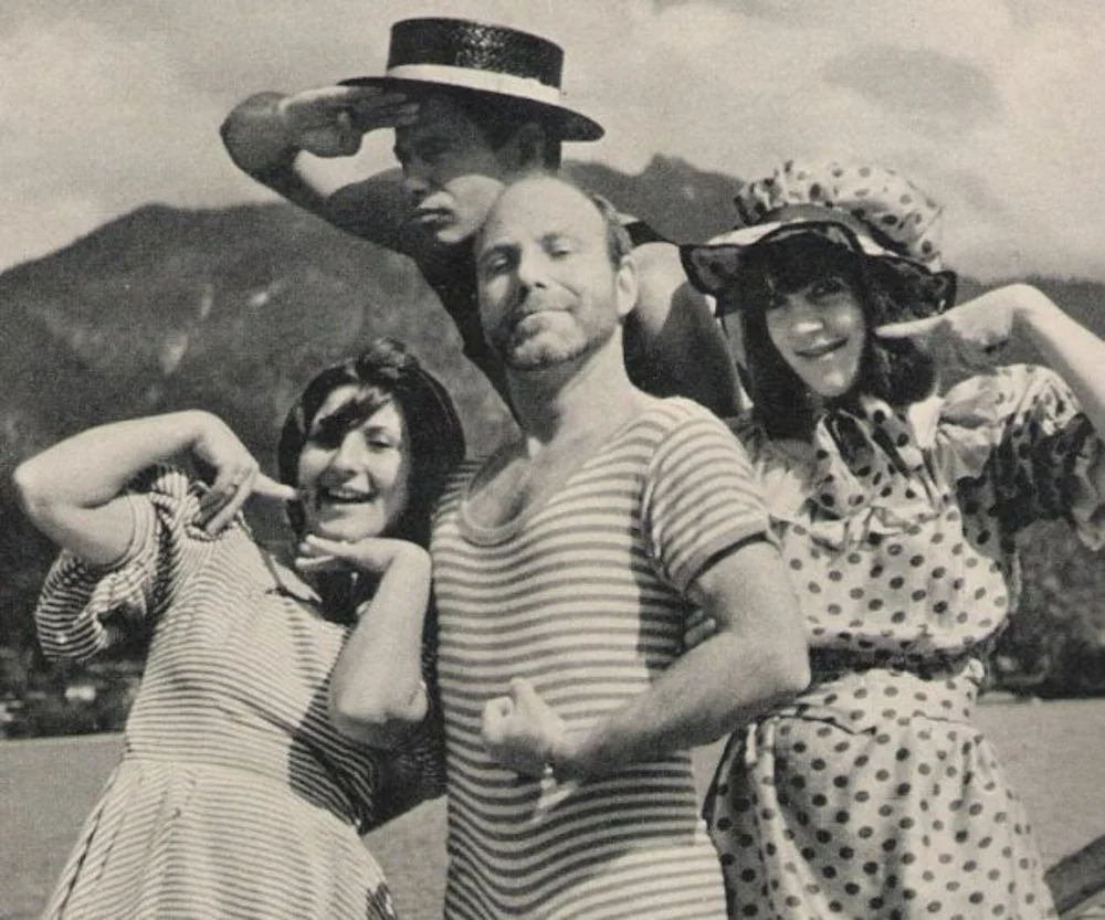 A black and white photo of four people outdoors, with mountains in the background. Two women are dressed in polka dot outfits, one with a large hat, and a man in the center with a striped shirt showing a flexed muscle. A young man behind them, wearin