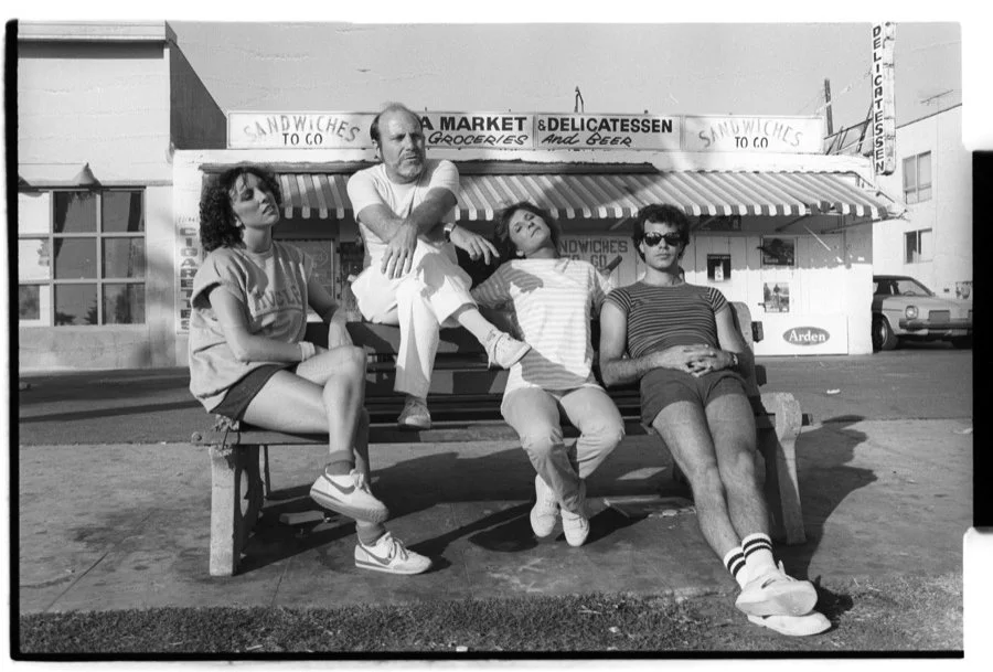 Four young adults sitting on a bench in front of a small grocery store with signs for sandwiches and delicatessen, some wearing casual clothes and sunglasses.