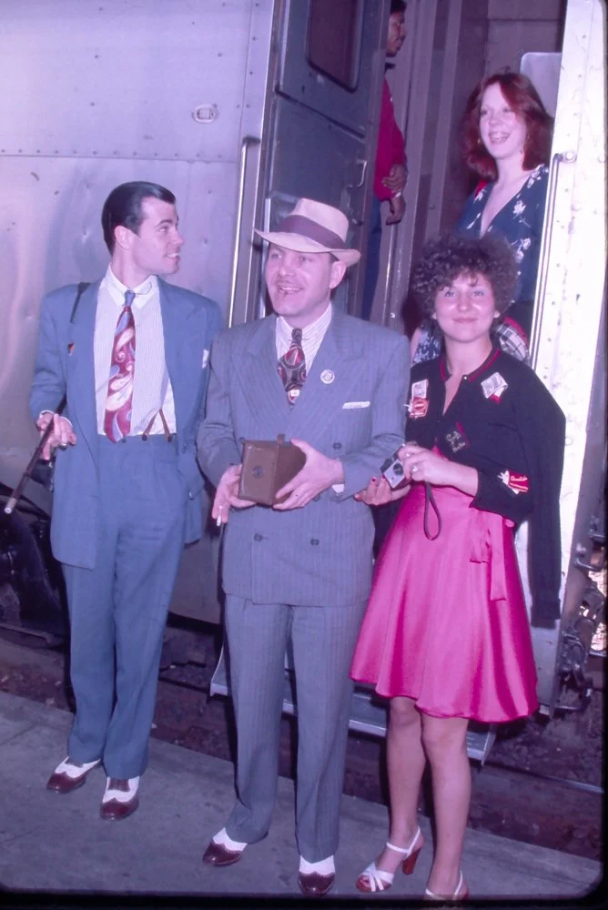 Four people posing outside a train or railcar. The man in the middle is wearing a gray pinstripe suit with a hat and holds a vintage camera. The woman on the right is dressed in a pink skirt with a black top and is holding a camera. The man on the le