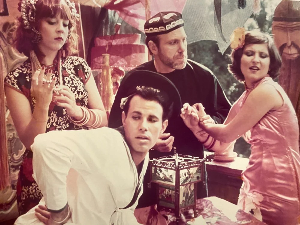 Four people dressed in traditional Chinese and Asian-inspired costumes participating in a role-playing scene. One woman in a cheongsam with floral headband, a man with a black hat and shirt, another woman in a pink satin dress, and a man in white with a black collar. They are indoors with ornate decorations and a large carved mask in the background.
