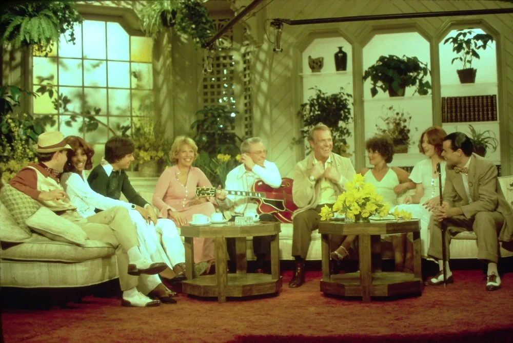 A group of nine people sitting on a beige couch and chairs, smiling and laughing, in a cozy living room with large windows, green plants, and bookshelves, with a woman playing an acoustic guitar.