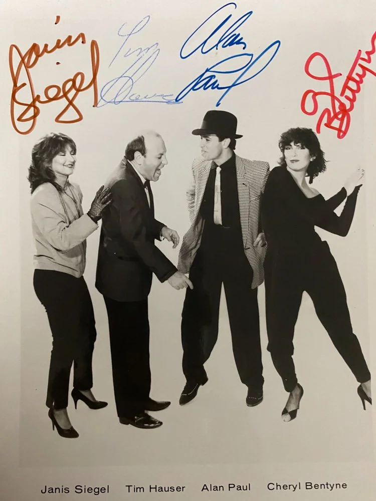 Black and white photograph of four people standing together, with signatures at the top. From left to right: Janis Siegel, Tim Hauser, Alan Paul, and Cheryl Bentyne.