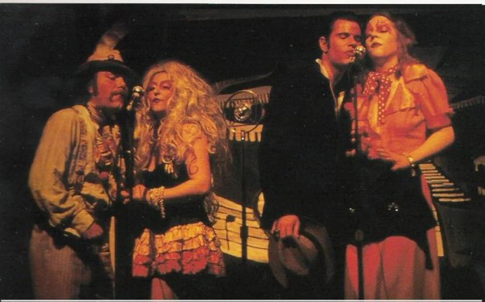 Four people dressed in vintage costumes stand on stage, with a keyboard in the background, performing or posing for a photo.