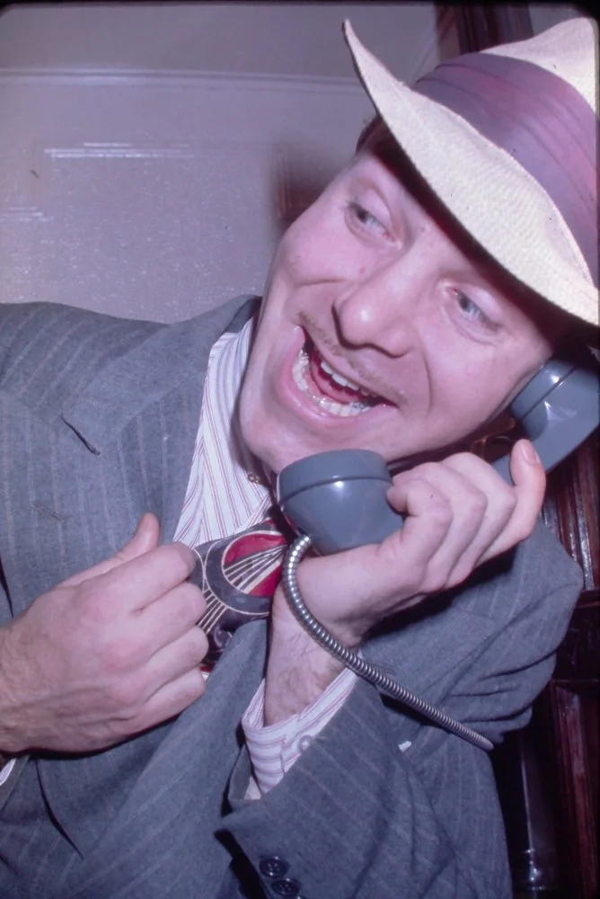 A man in a suit and striped shirt, wearing a fedora, smiling and speaking on a landline phone, with a lively expression.