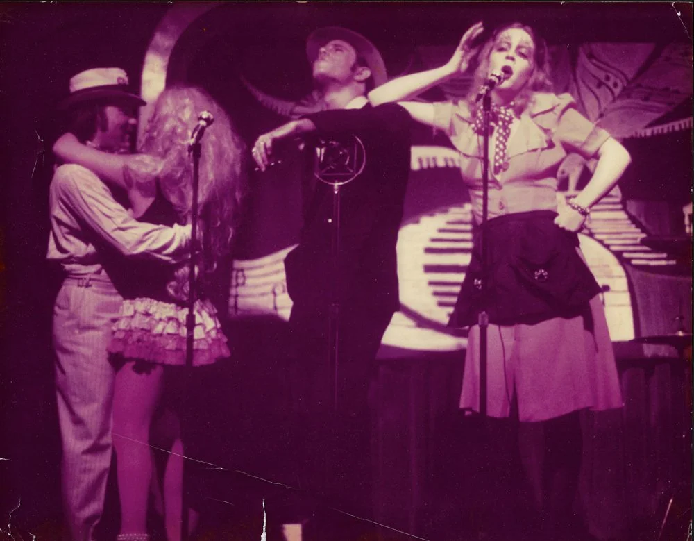 Four performers on stage, two women and two men, with a background of a painted piano and musical notes. One woman wears a hat and striped outfit, the other wears a skirt and blouse, the men wear casual clothing and hats, engaged in singing or acting