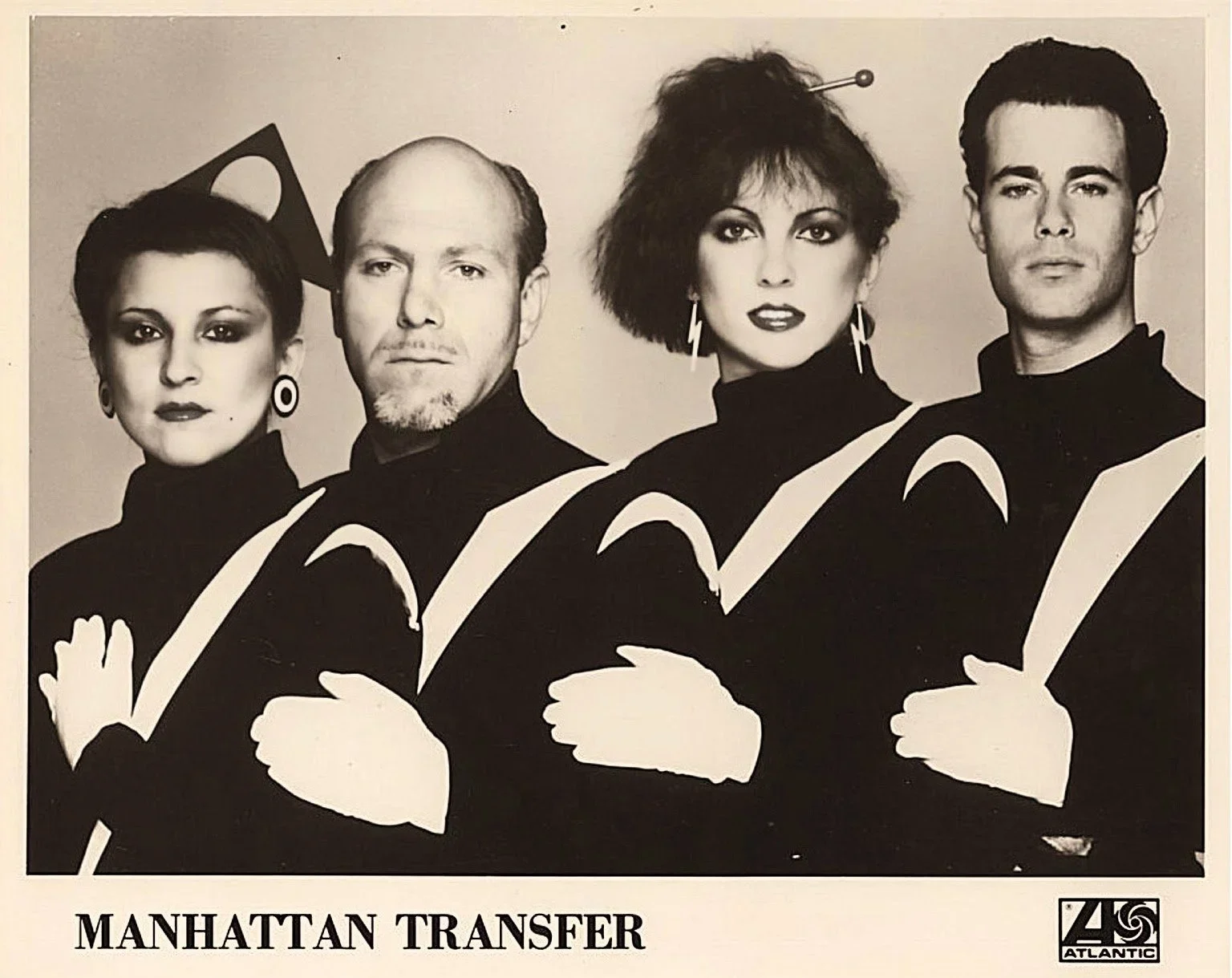 Black and white promotional photo of four members of the band Manhattan Transfer, dressed in coordinated black outfits with white stripes, standing with arms crossed. The band's name is written below the photo along with the Atlantic Records logo.