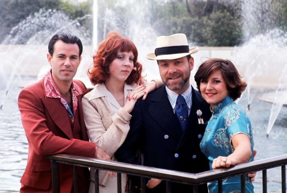 Four people standing together on a boat with a fountain and water in the background.