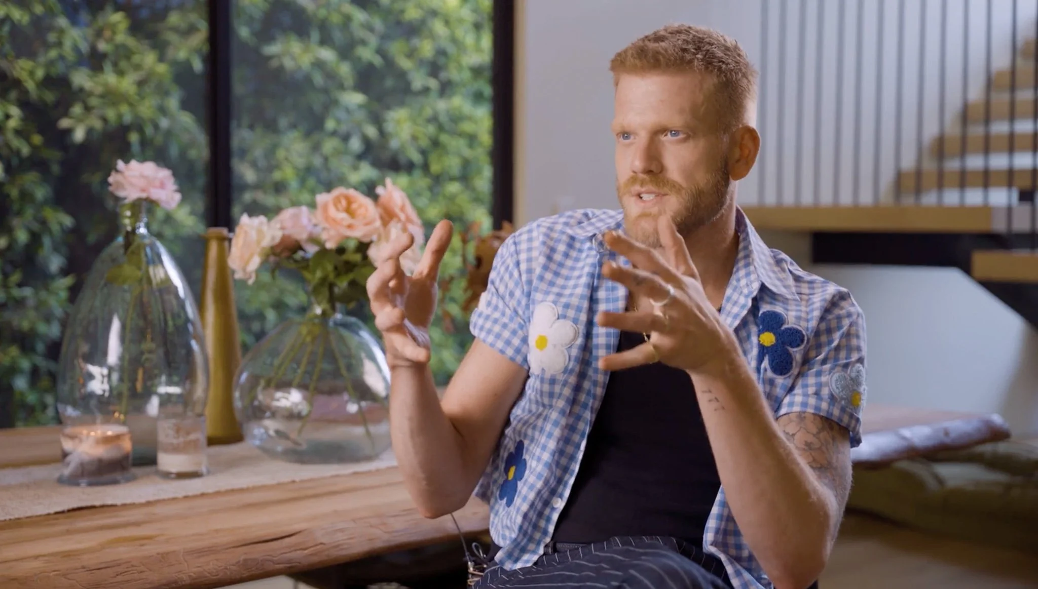 A man with short red hair, a beard, and tattoos on his arm, wearing a blue and white checkered shirt with floral patches, sitting at a wooden table indoors, gesturing with his hands while speaking. Behind him is a large window with a view of green foliage, and on the table are decorative vases with pink flowers and candles.